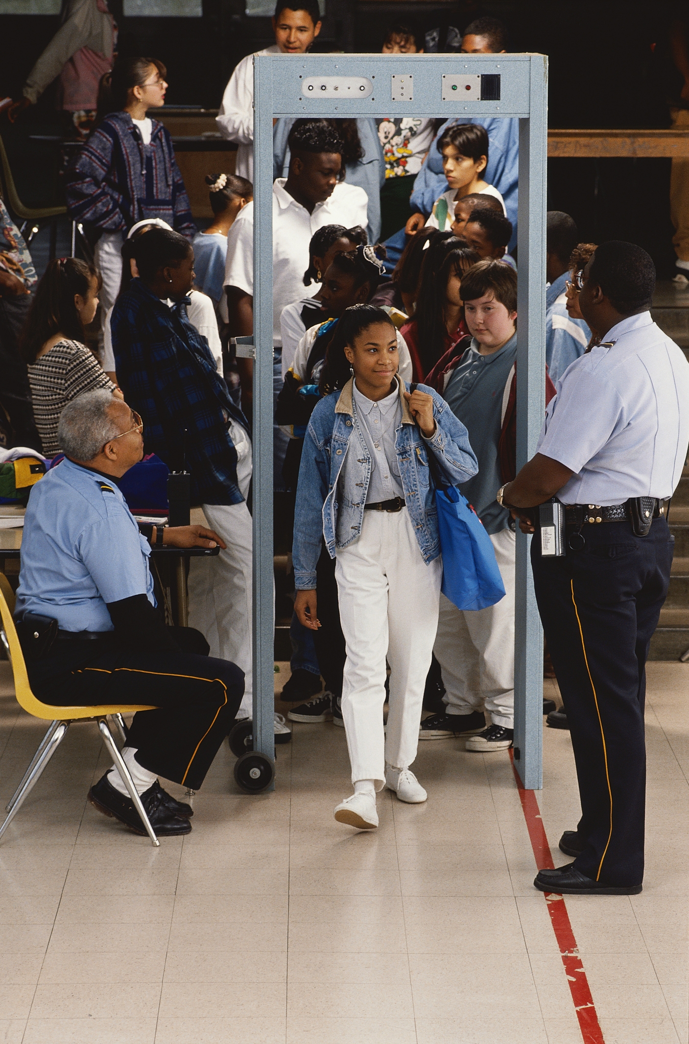 A large group of school students pass through a metal detector monitored by security guards indoors