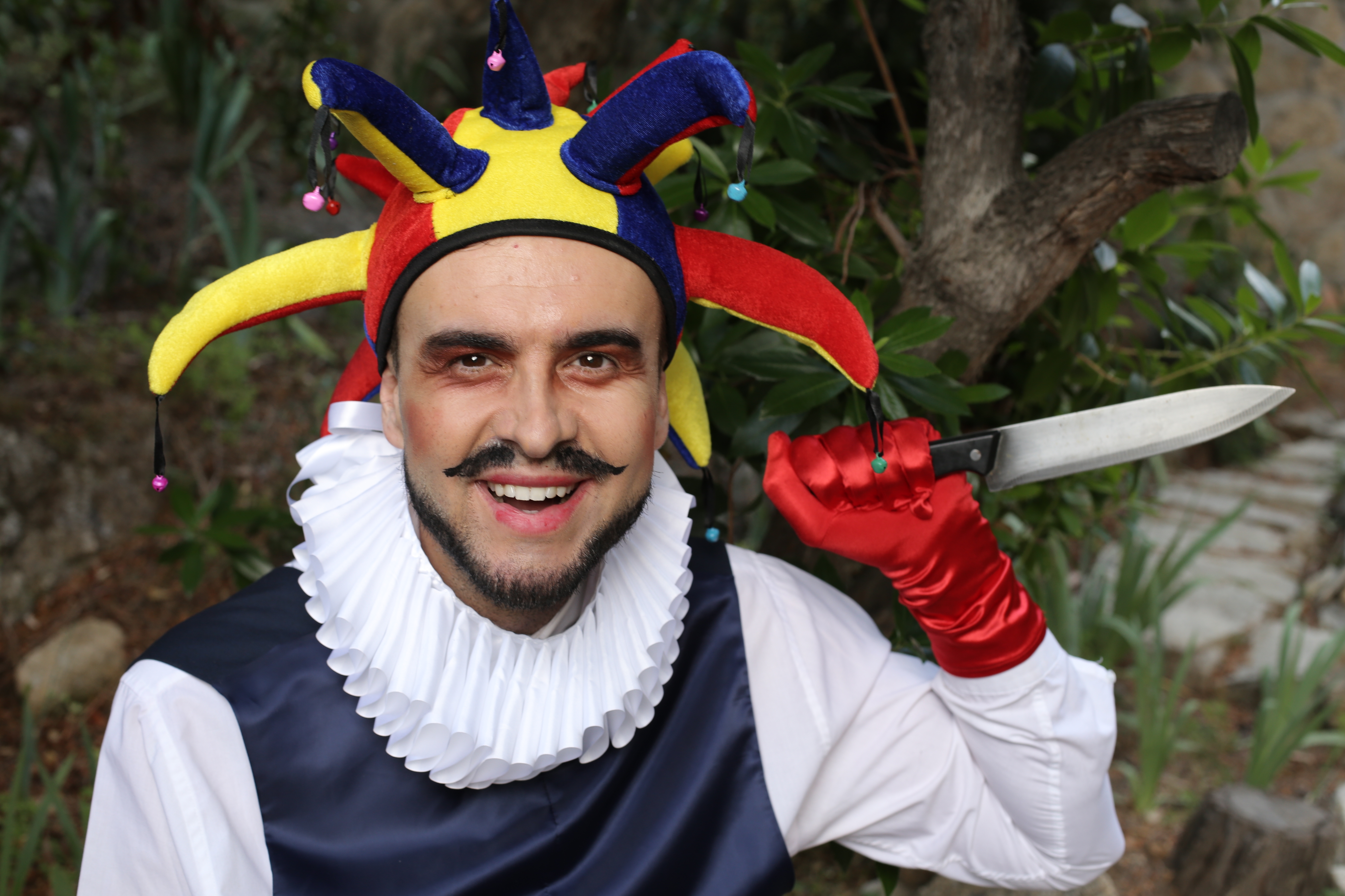 Person in jester costume with a ruffled collar, wearing a playful hat, holding a theatrical knife, smiling in a garden setting