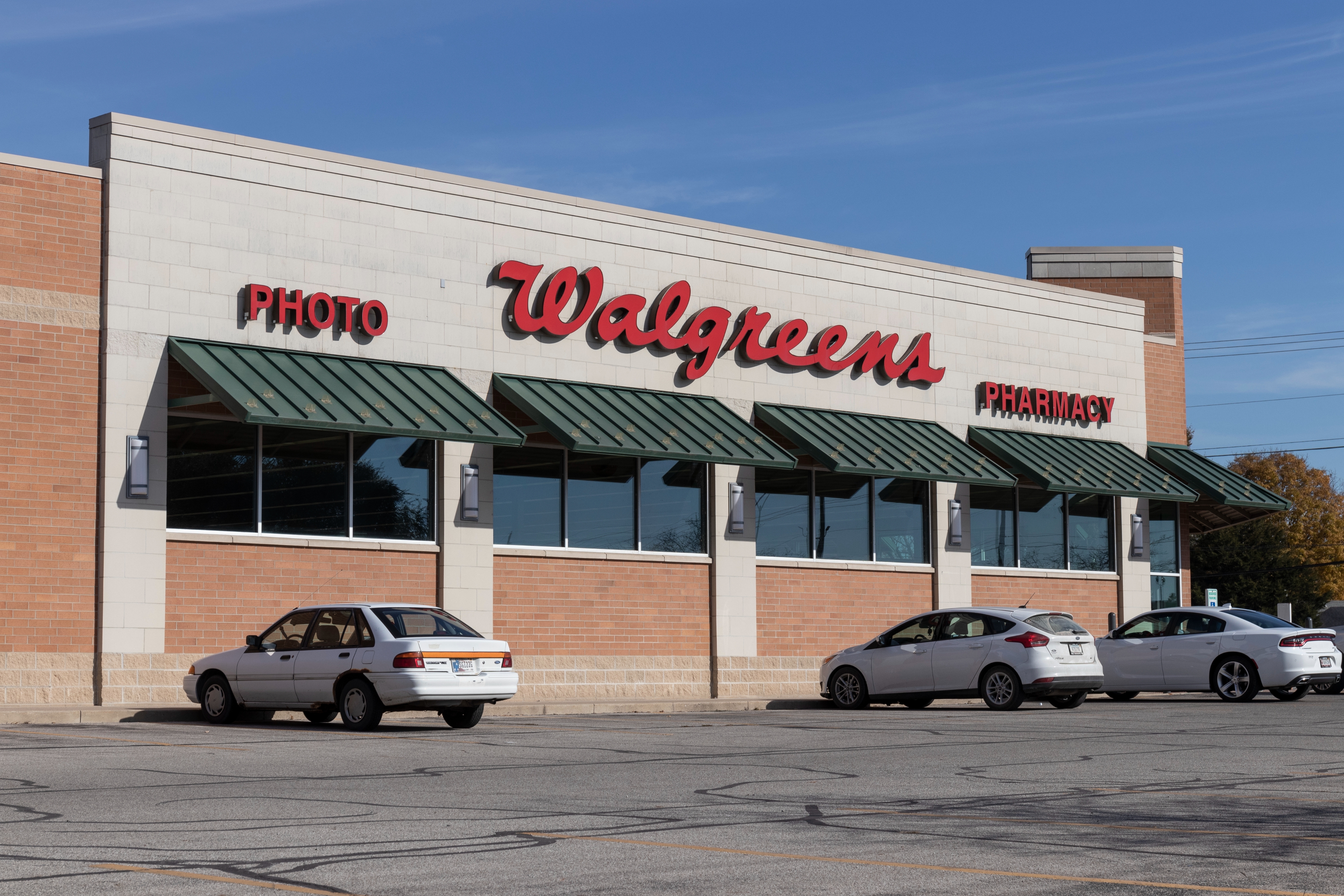 Exterior of a Walgreens pharmacy with several parked cars in the lot. The building has signage for "Photo" and "Pharmacy."