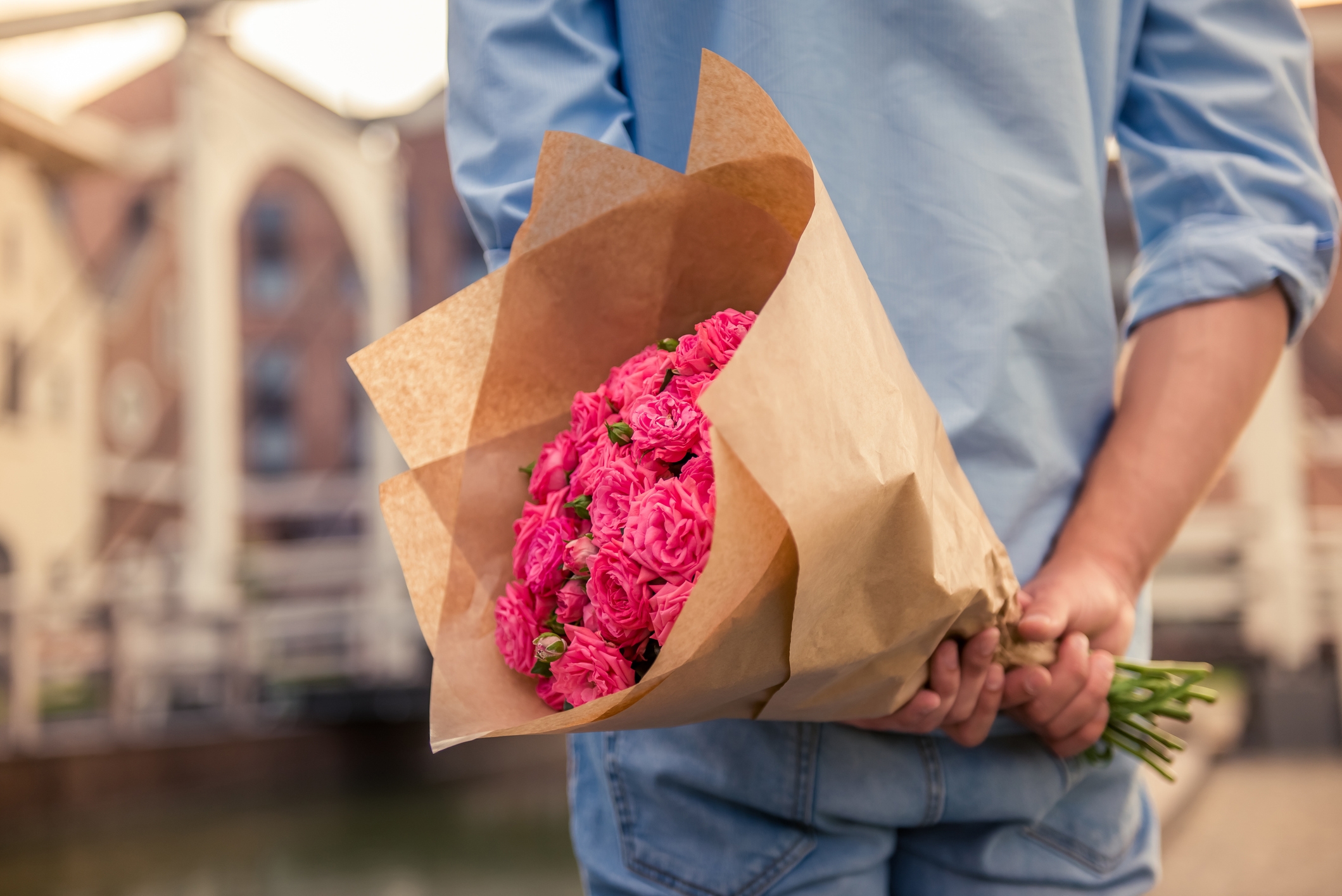 Person holding a bouquet of pink flowers wrapped in brown paper behind their back, standing outside