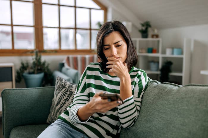 A person sits on a couch, looking intently at their phone, wearing a striped top. There are plants and shelves in the background