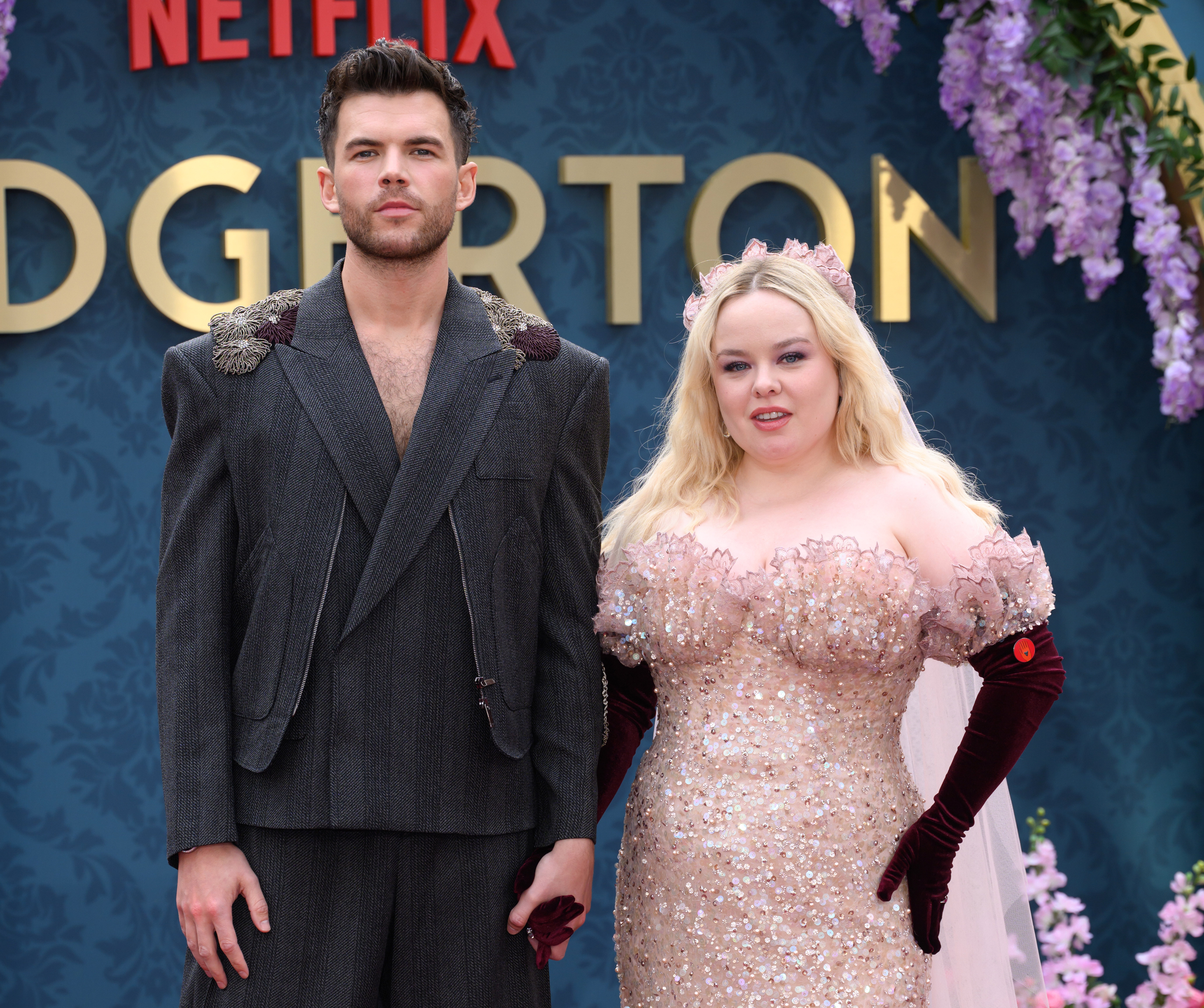 Two people on a red carpet; one wears a dark, structured suit, and the other an embellished dress with floral details