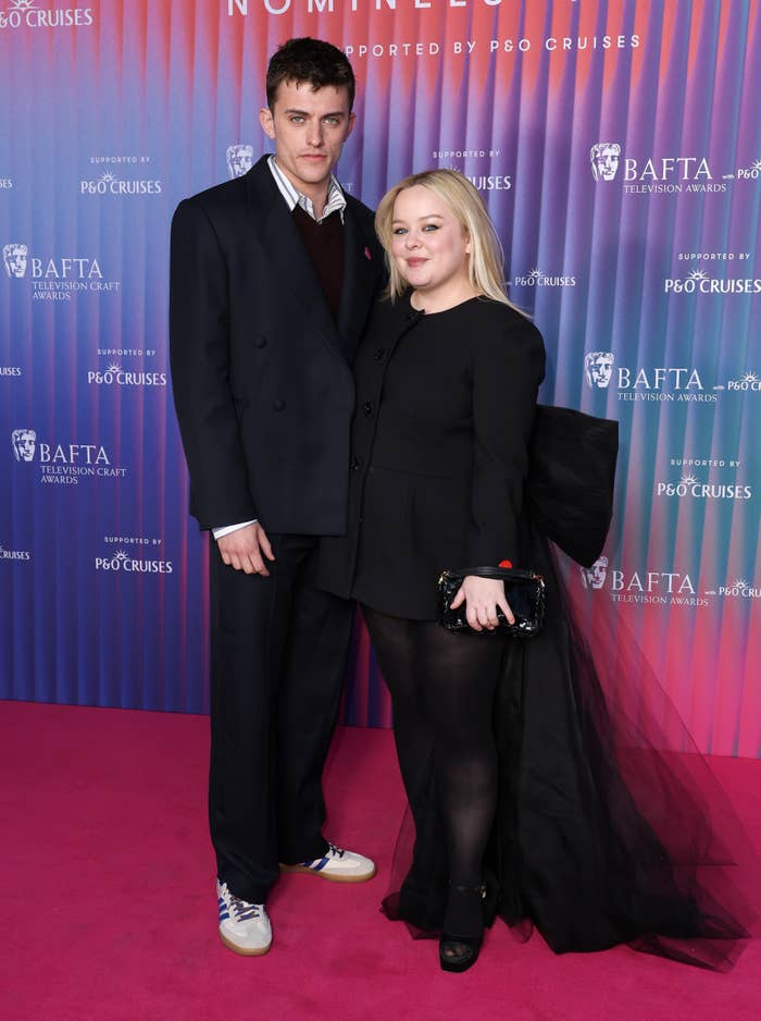 Two individuals stand on a red carpet. One wears a dark suit with sneakers, and the other is in a stylish black dress