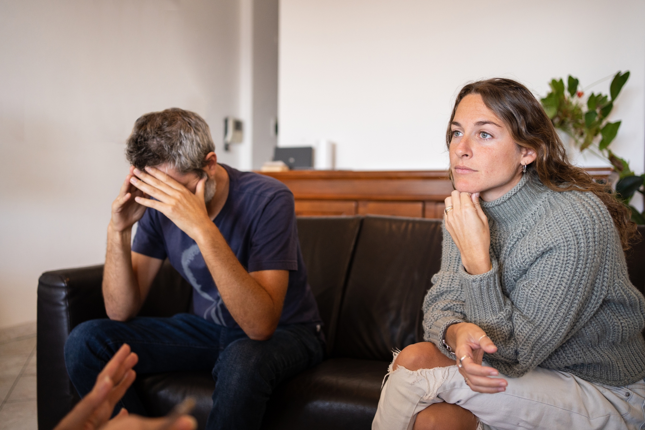 A woman in a sweater sits pensively on a couch, while a man beside her covers his face with his hands. A third person gestures during a conversation