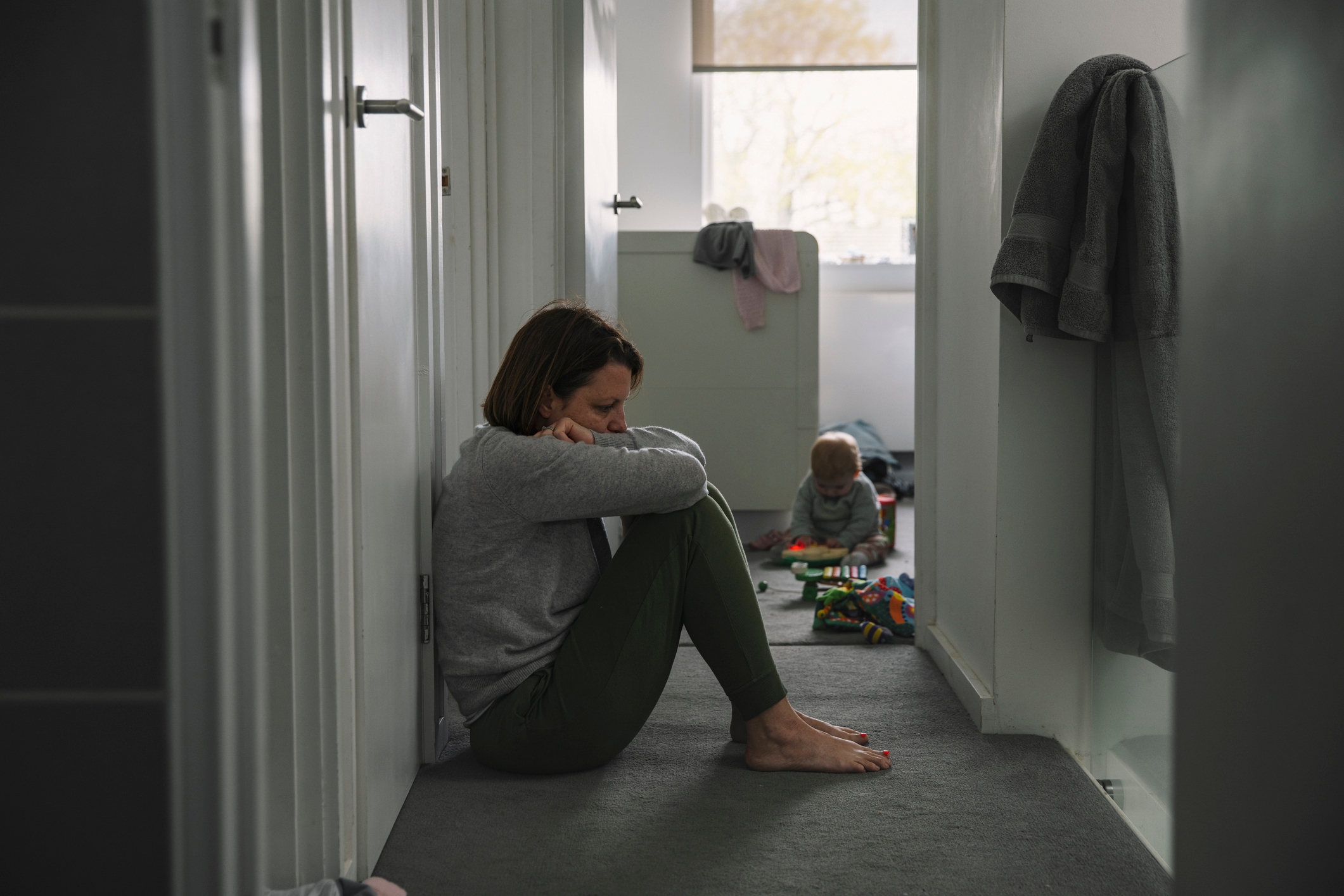 Person sitting in a hallway with a thoughtful expression; a child is playing with toys in the background