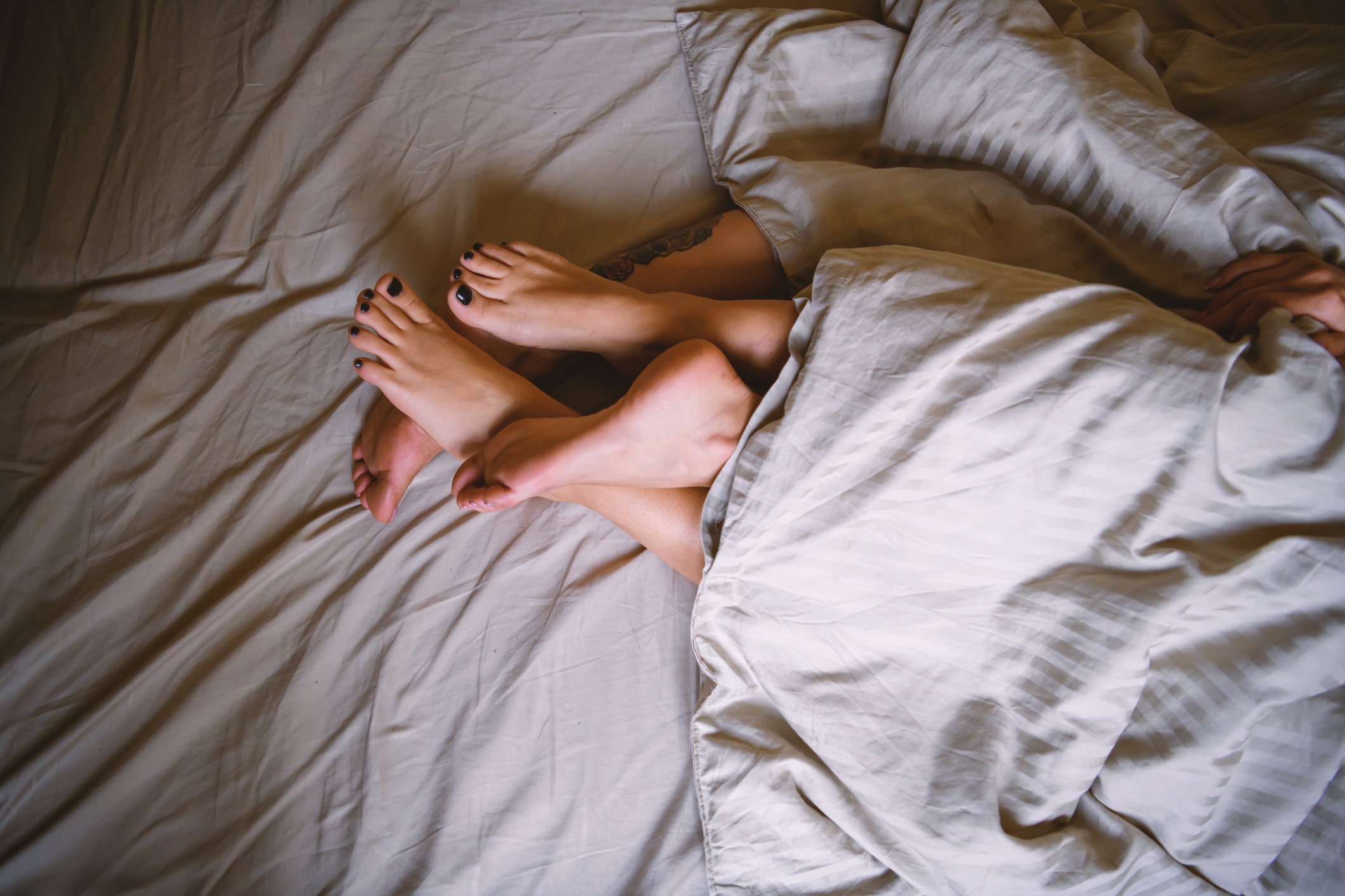 Two pairs of adult feet are intertwined under a blanket on a bed