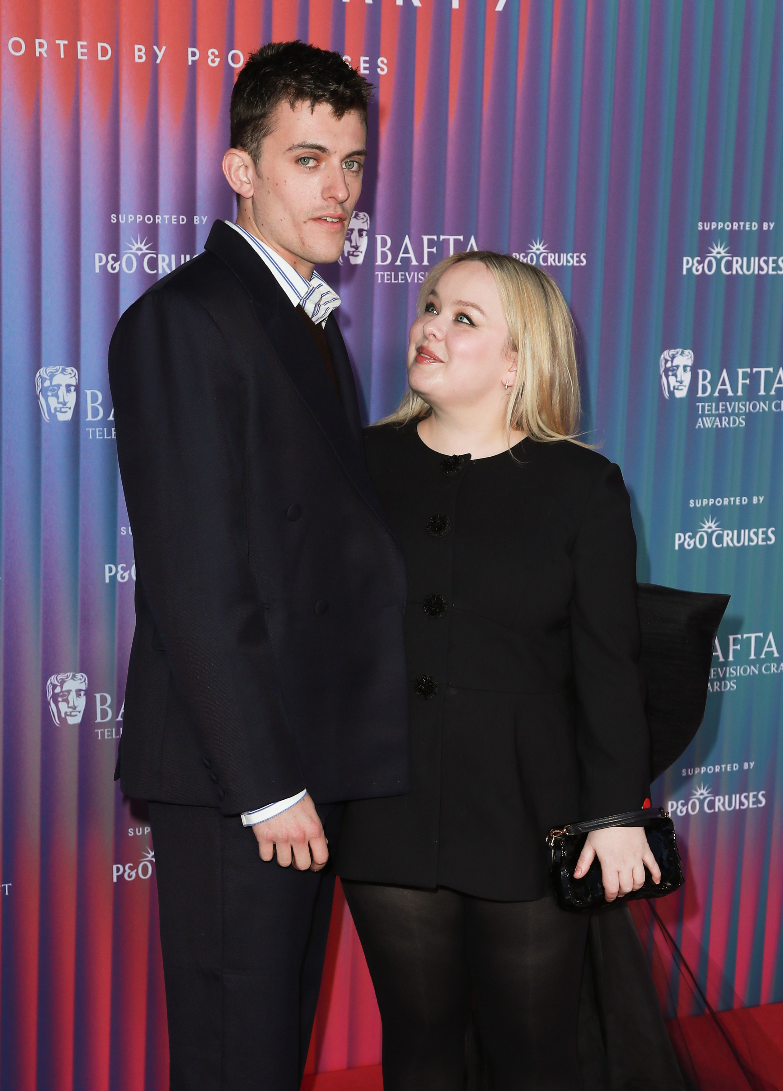 Two people at a BAFTA event. The man wears a dark suit, and the woman wears a black outfit, holding a clutch. They pose in front of a promotional backdrop
