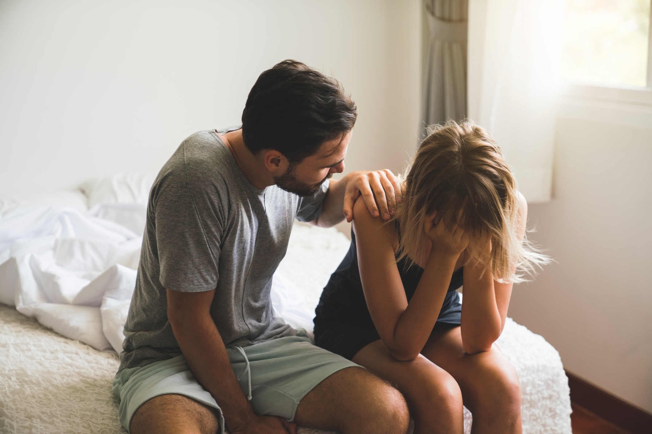 A man in casual clothes sits on a bed, comforting a woman who is seated and holding her head in distress