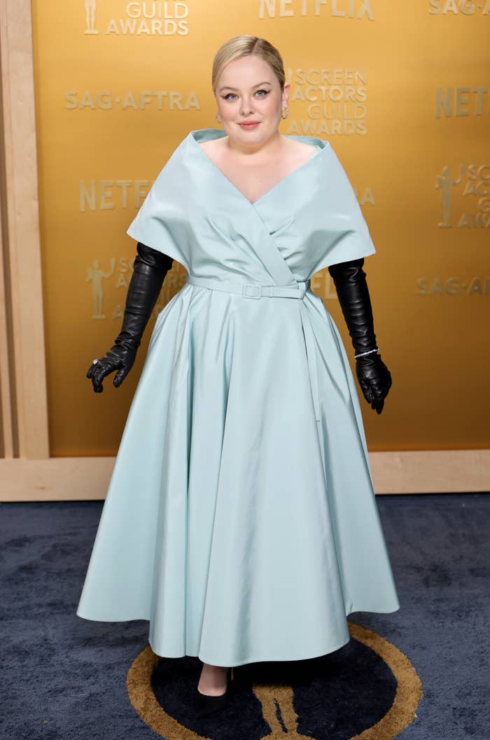 Person in a stylish gown with a fitted waist and wide collar, wearing long gloves, posing on a red carpet at an award event