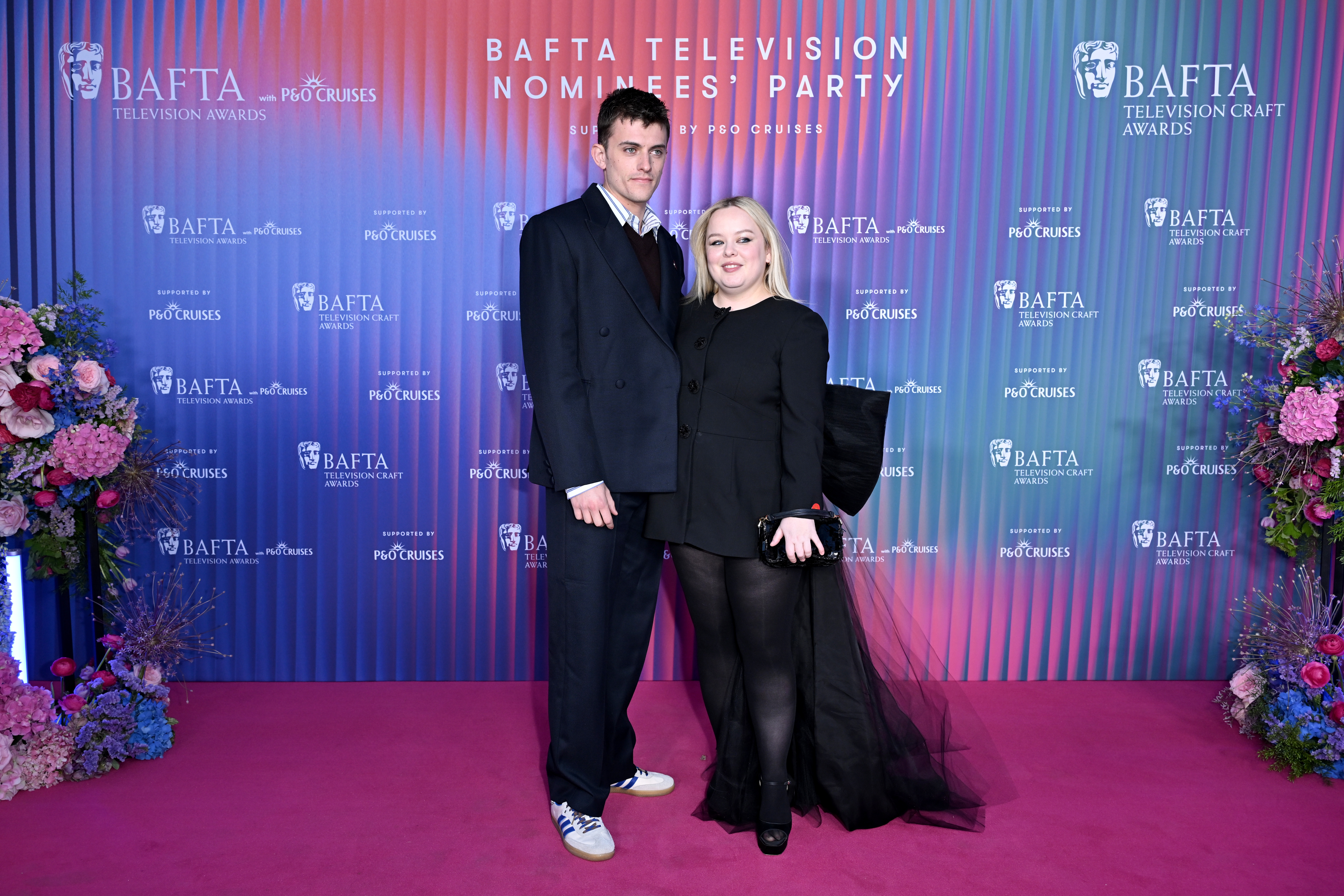 Two individuals pose on a BAFTA event red carpet. One wears a suit; the other wears a chic black outfit with a tulle train