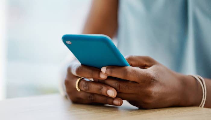 Person holding a smartphone with both hands, resting elbows on a table. They wear rings and a bracelet, focusing on the screen