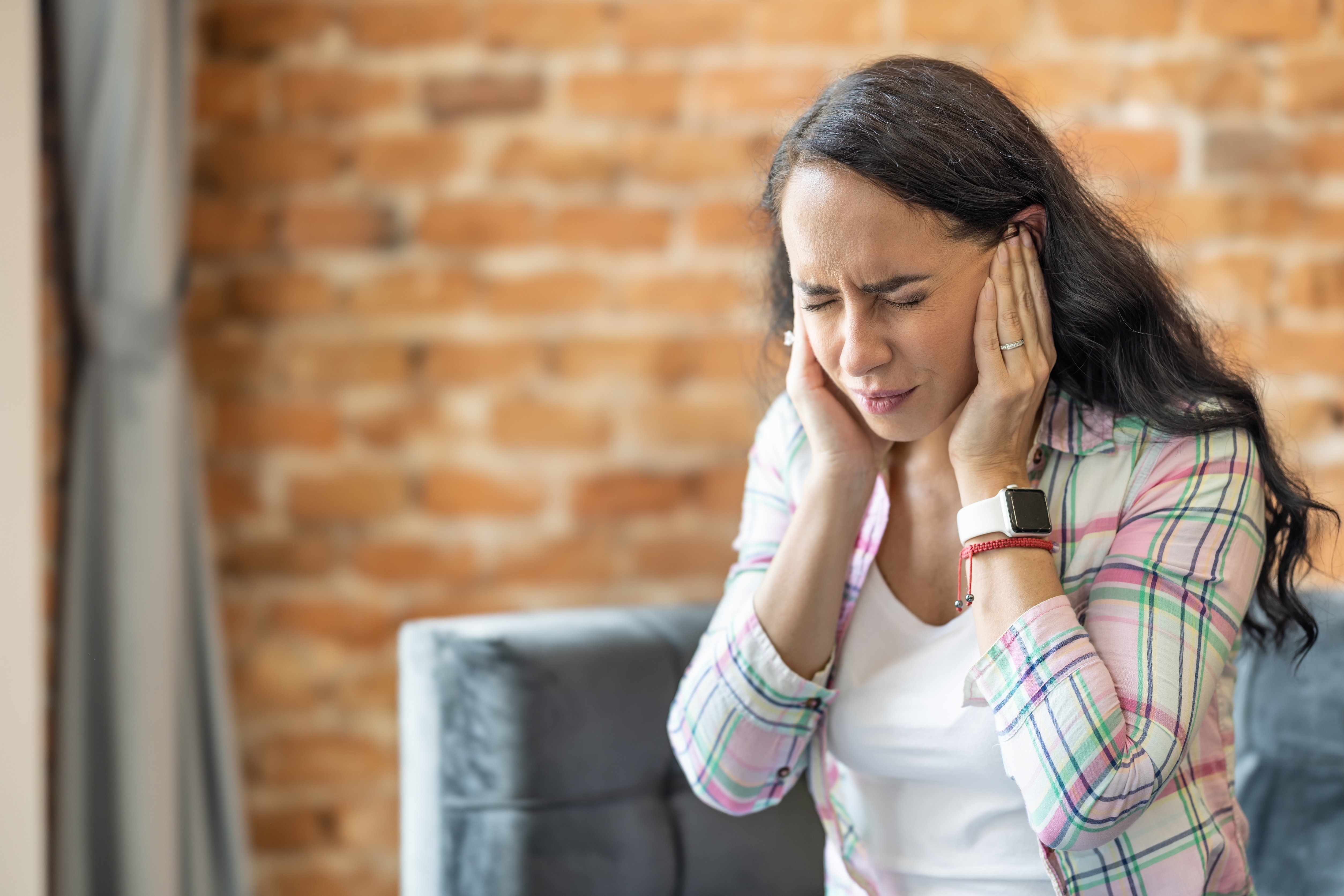 A woman sitting on a couch holds her head with both hands, appearing stressed or in discomfort, in a brick-walled room