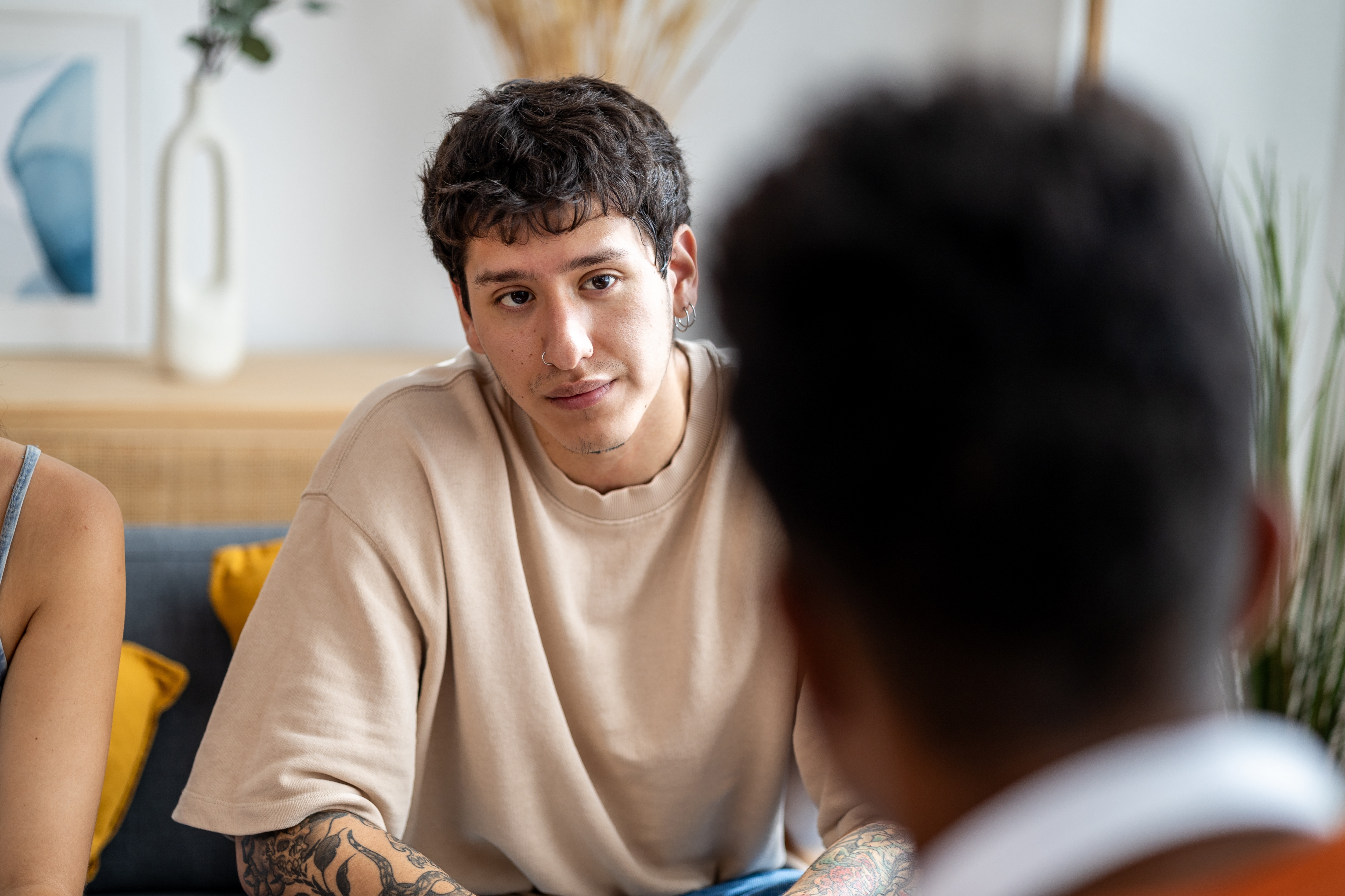 A person with tattoos and a casual shirt attentively listens to someone in a casual indoor setting