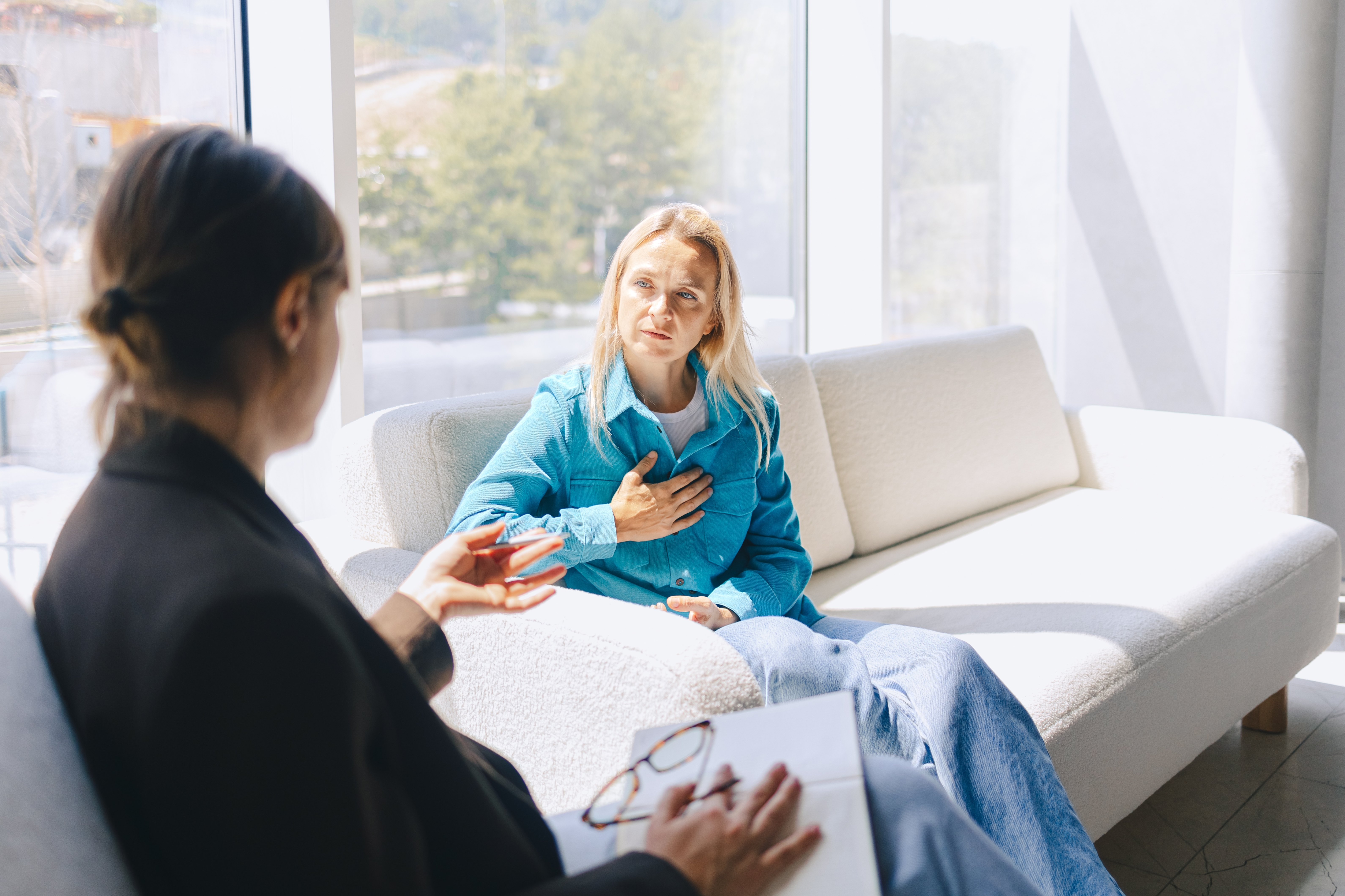 Two people are seated on a couch in conversation. One person gestures with their hand to their chest, while the other holds a clipboard