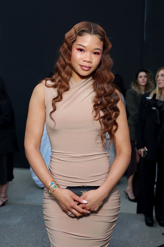 Person poses in a form-fitting sleeveless dress at a celebrity event, holding a clutch. Wavy hair and vibrant eye makeup accentuate the look
