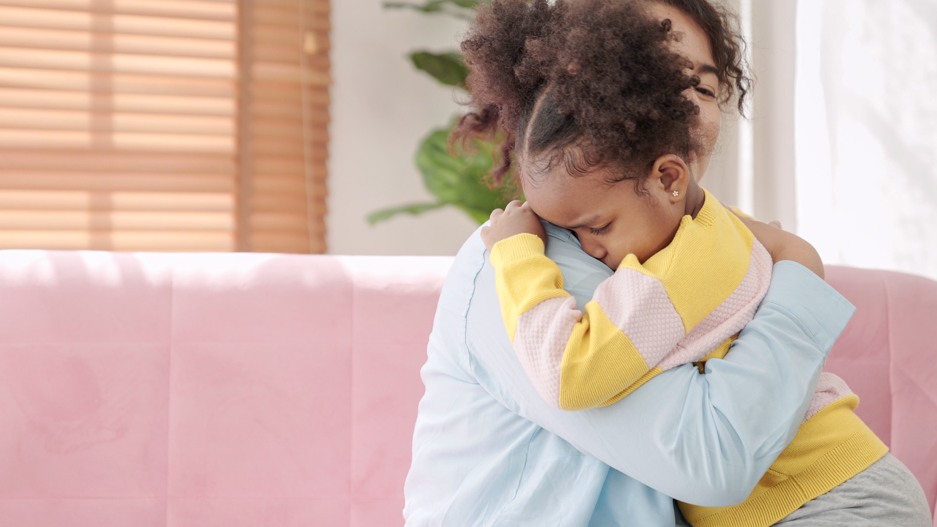 A child with curly hair and a colorful sweater is being comforted in an embrace by an adult on a couch, indicating a moment of tenderness