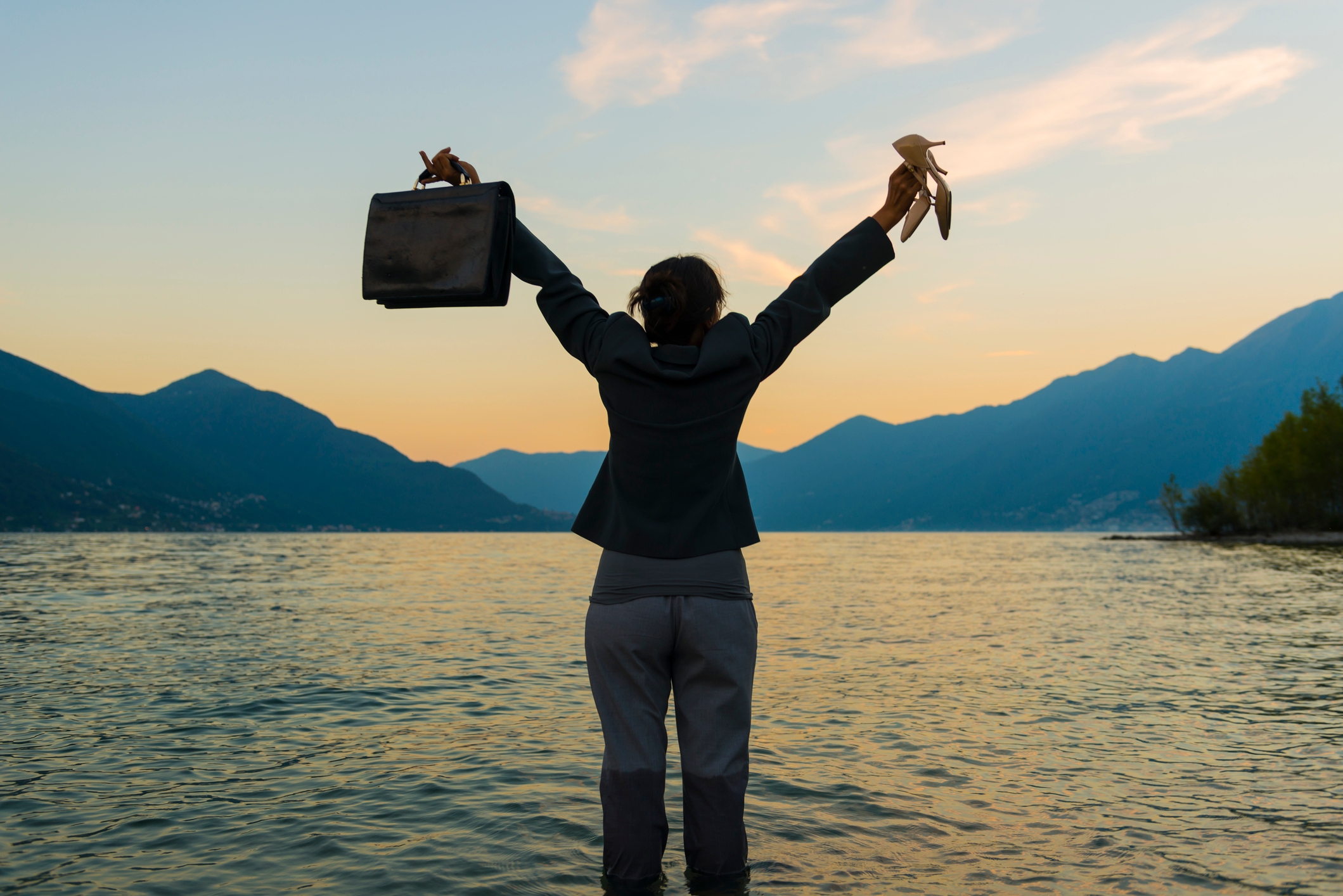 Person standing in water holding shoes and briefcase, arms raised in triumph, with mountains in the distance and a serene sunset sky