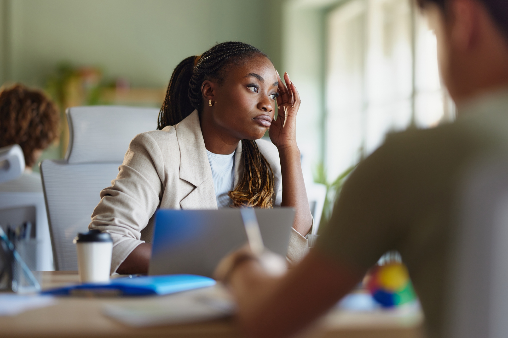 Woman in office setting looking pensive, resting head on hand, laptop open in front of her, suggesting work-related contemplation