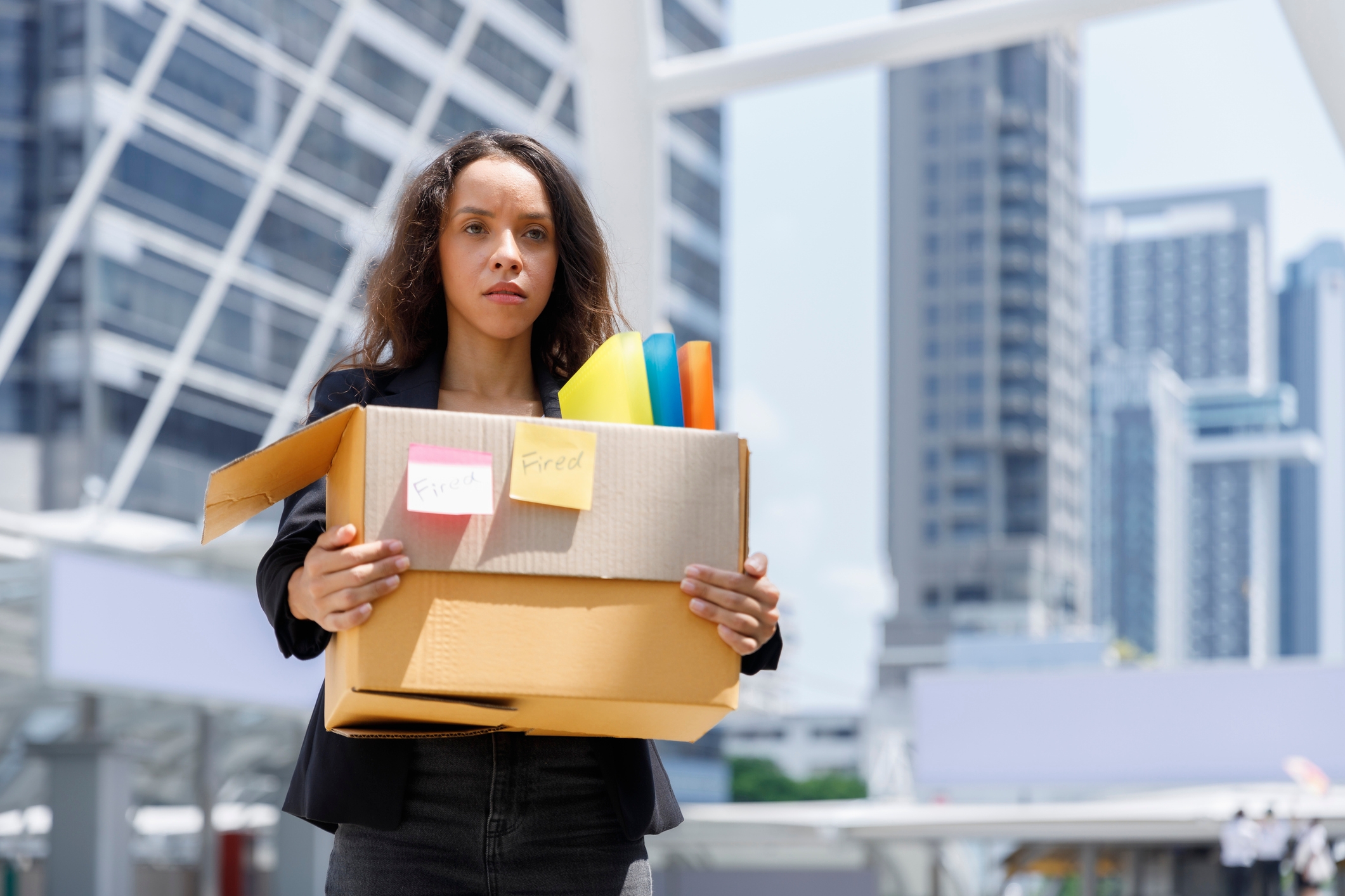 Woman in business attire holding a cardboard box with office items, standing in an urban setting, possibly symbolizing job loss or career transition