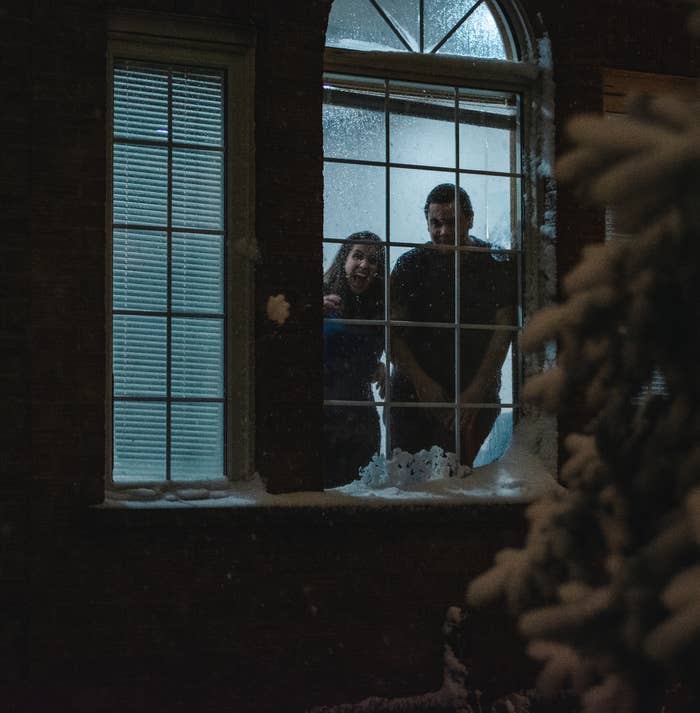 Two people smiling, looking out a snow-covered window at night, creating a cozy, festive atmosphere