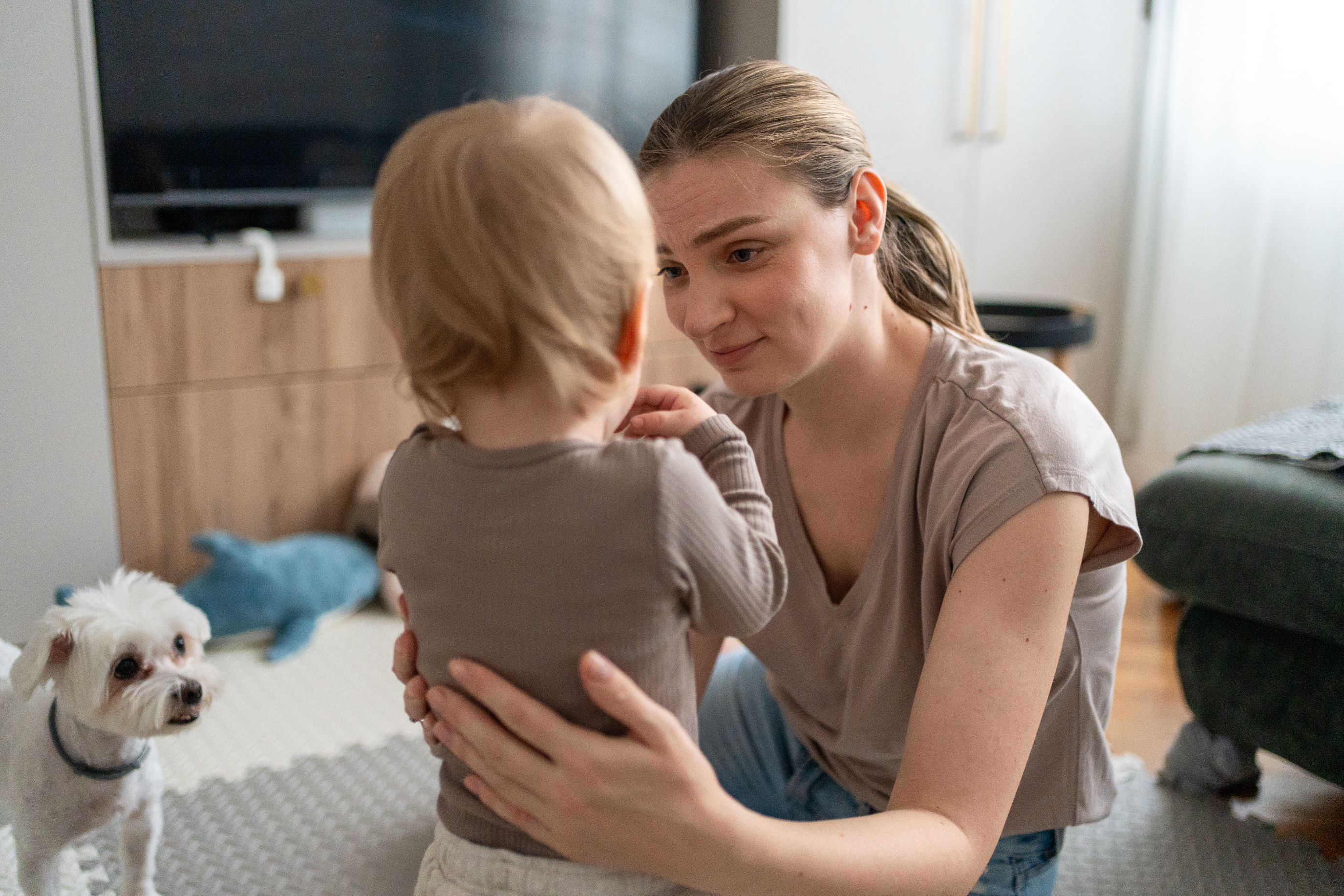 Woman kneeling, gently talking to toddler with a small white dog nearby in a living room setting