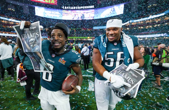 Two football players celebrate on the field holding championship newspapers, wearing jerseys and victory gear, surrounded by confetti