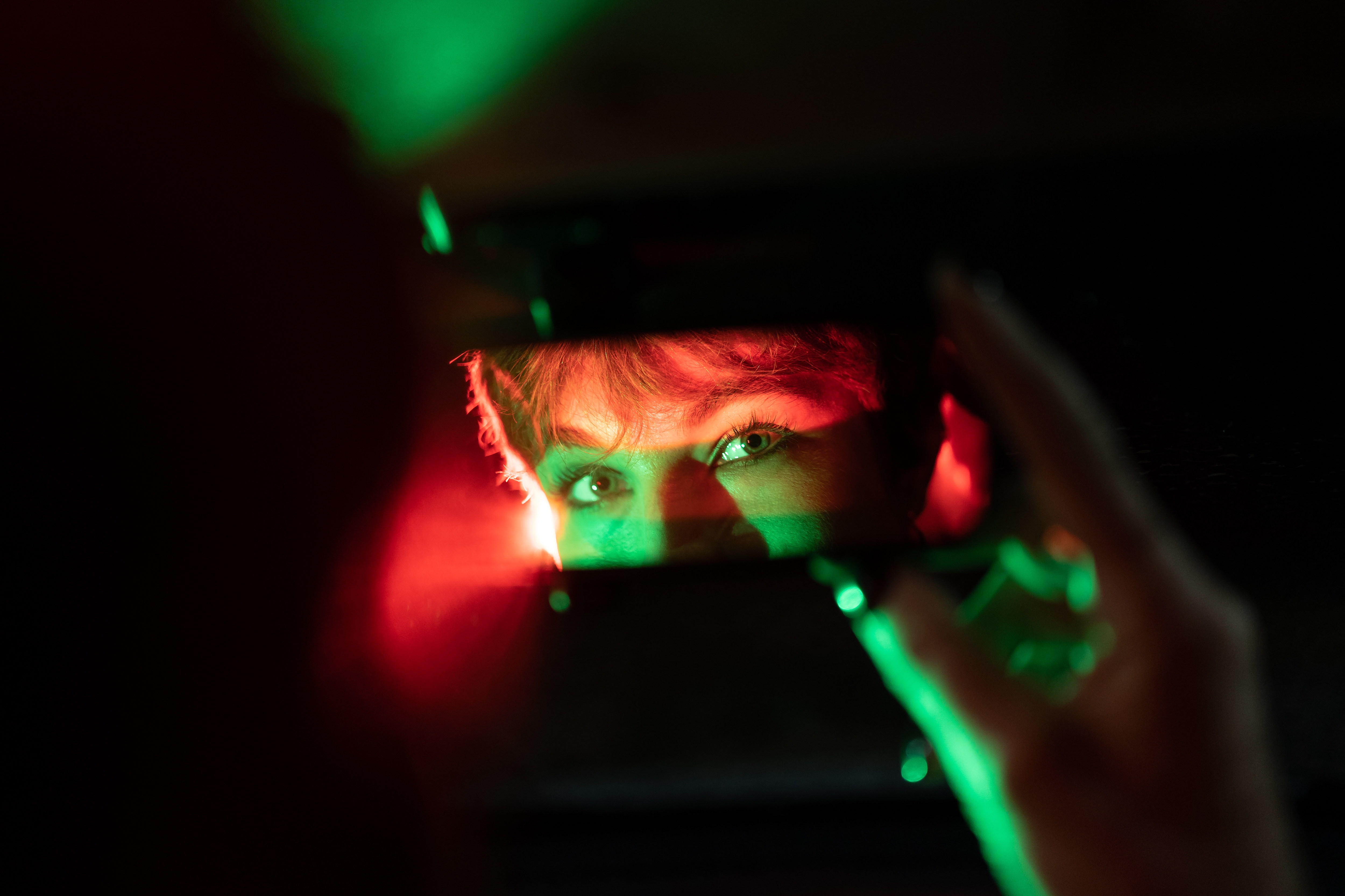 Person's eyes reflected in a small mirror, partially obscured by shadows and colored lights. Mysterious, introspective atmosphere