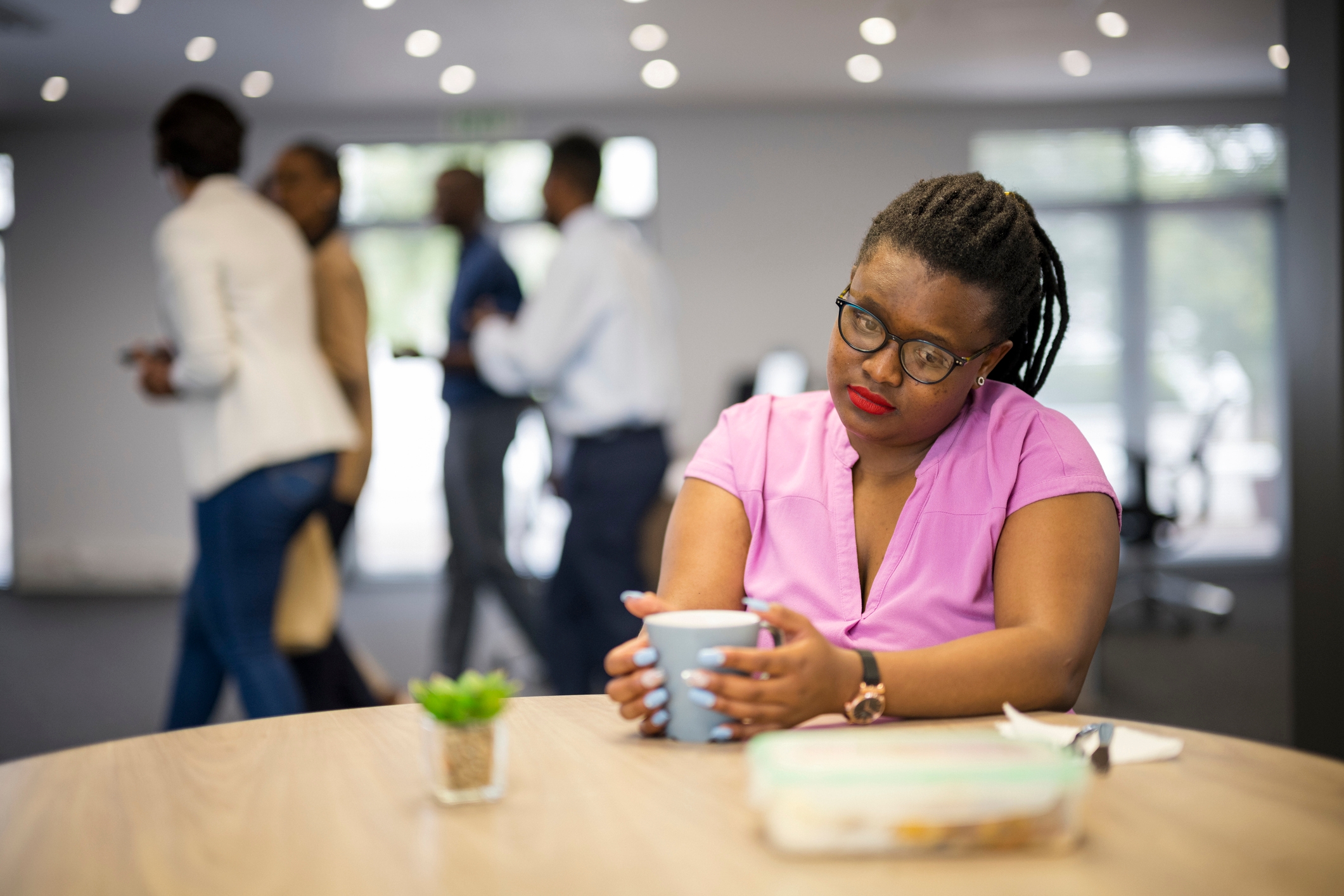 Person with glasses sits at a desk holding a coffee cup, looking thoughtful, while colleagues chat in the background. Office setting