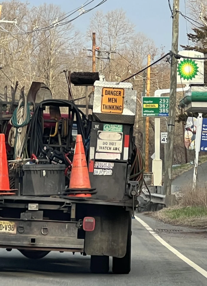 Truck with tools and signs speechmaking  "Danger Men Thinking" and "Do Not Watch Arc" connected  a roadworthy  adjacent   a state  station