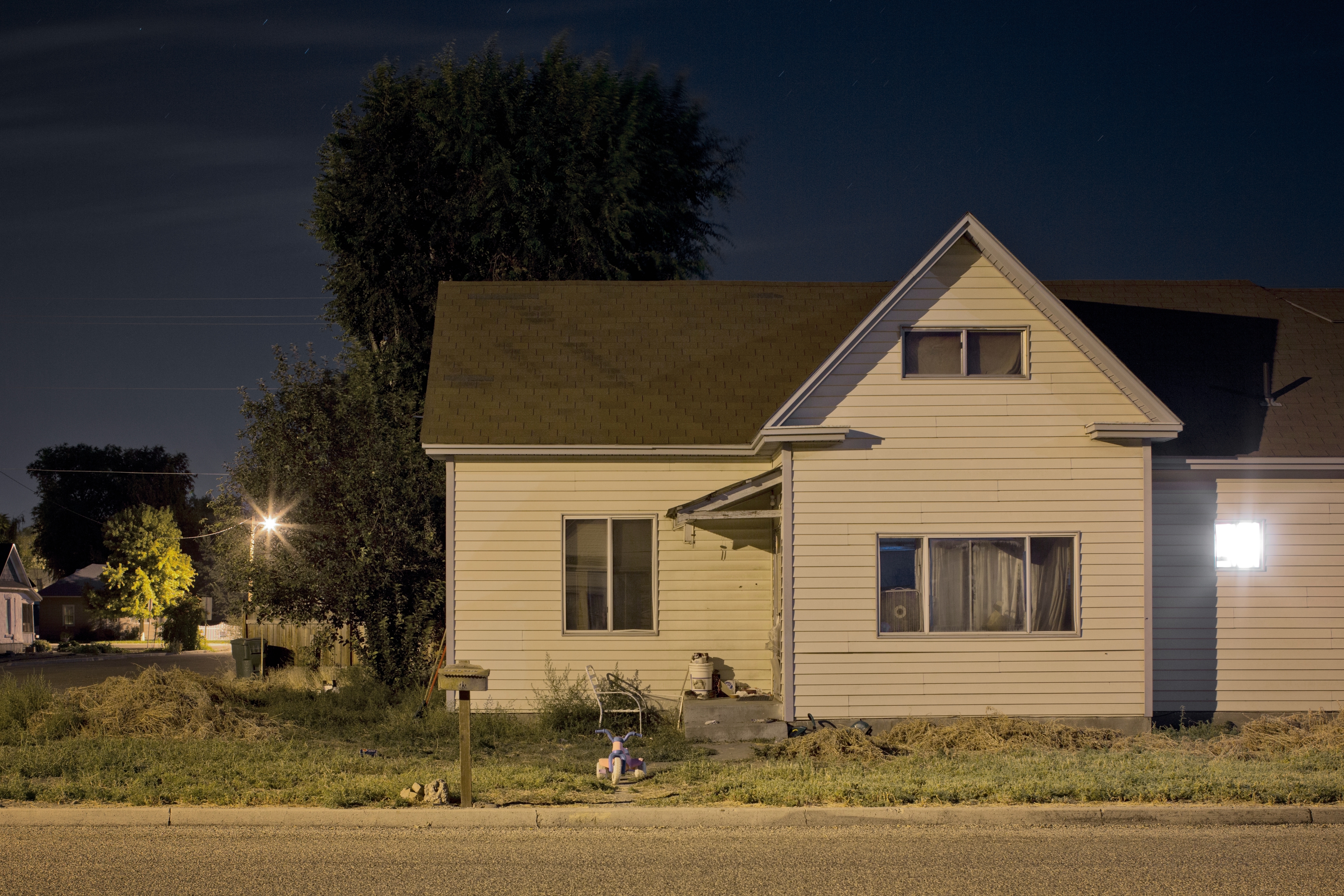 A small, single-story house at night with a tree nearby and a streetlamp illuminating the scene. A bicycle is visible on the porch