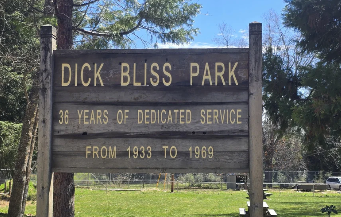 Wooden sign for Dick Bliss Park reads: "36 years of dedicated service from 1933 to 1969." Trees and grass surround the sign