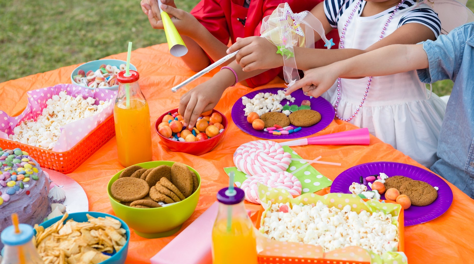 Children in costumes at an outdoor party table with snacks like popcorn, cookies, and candy