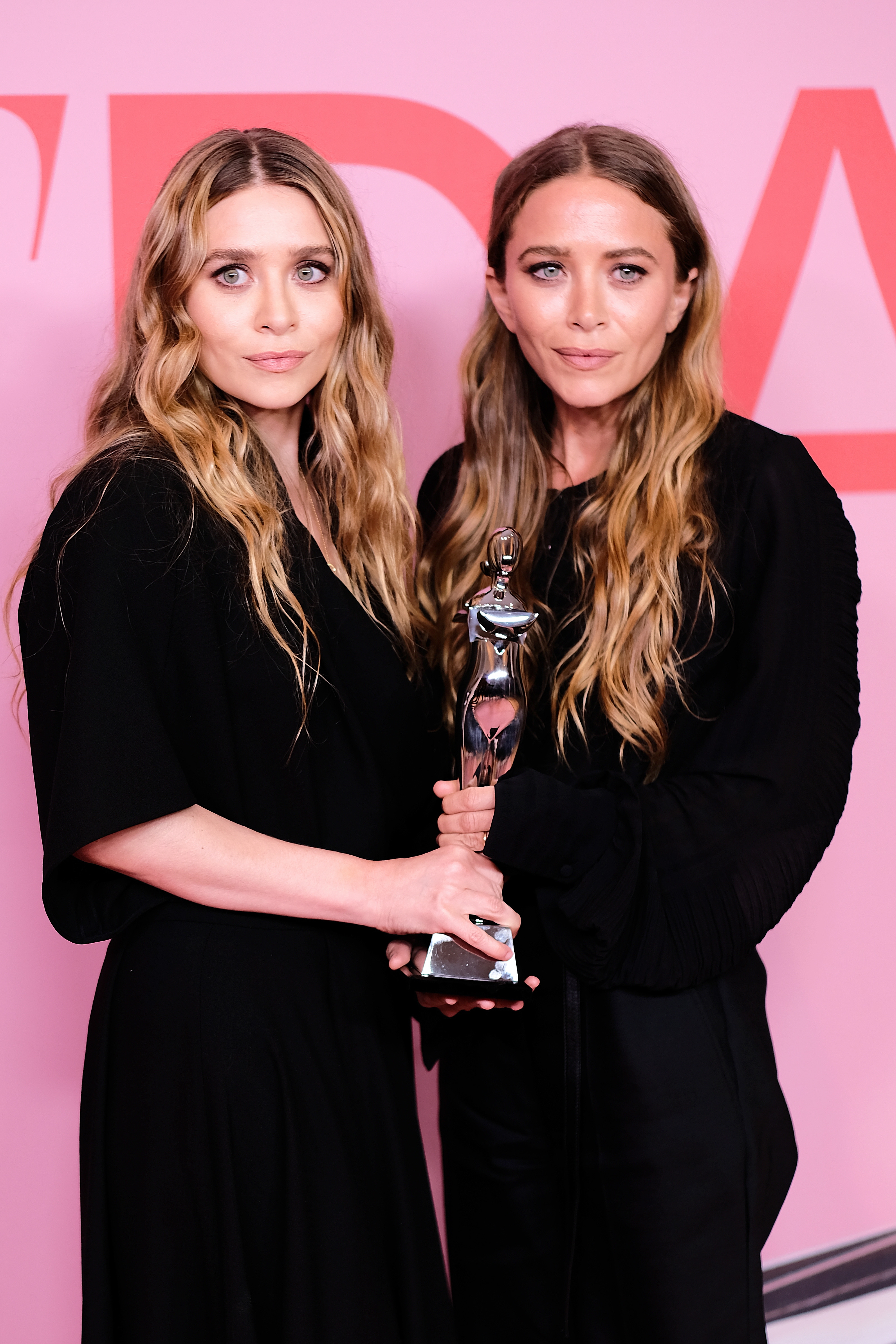 Two women with long, wavy hair in elegant black outfits holding an award statue at a formal event