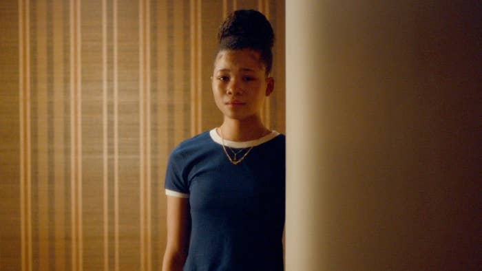 Person stands alone indoors, looking contemplative. They wear a simple, fitted short-sleeve top and a necklace