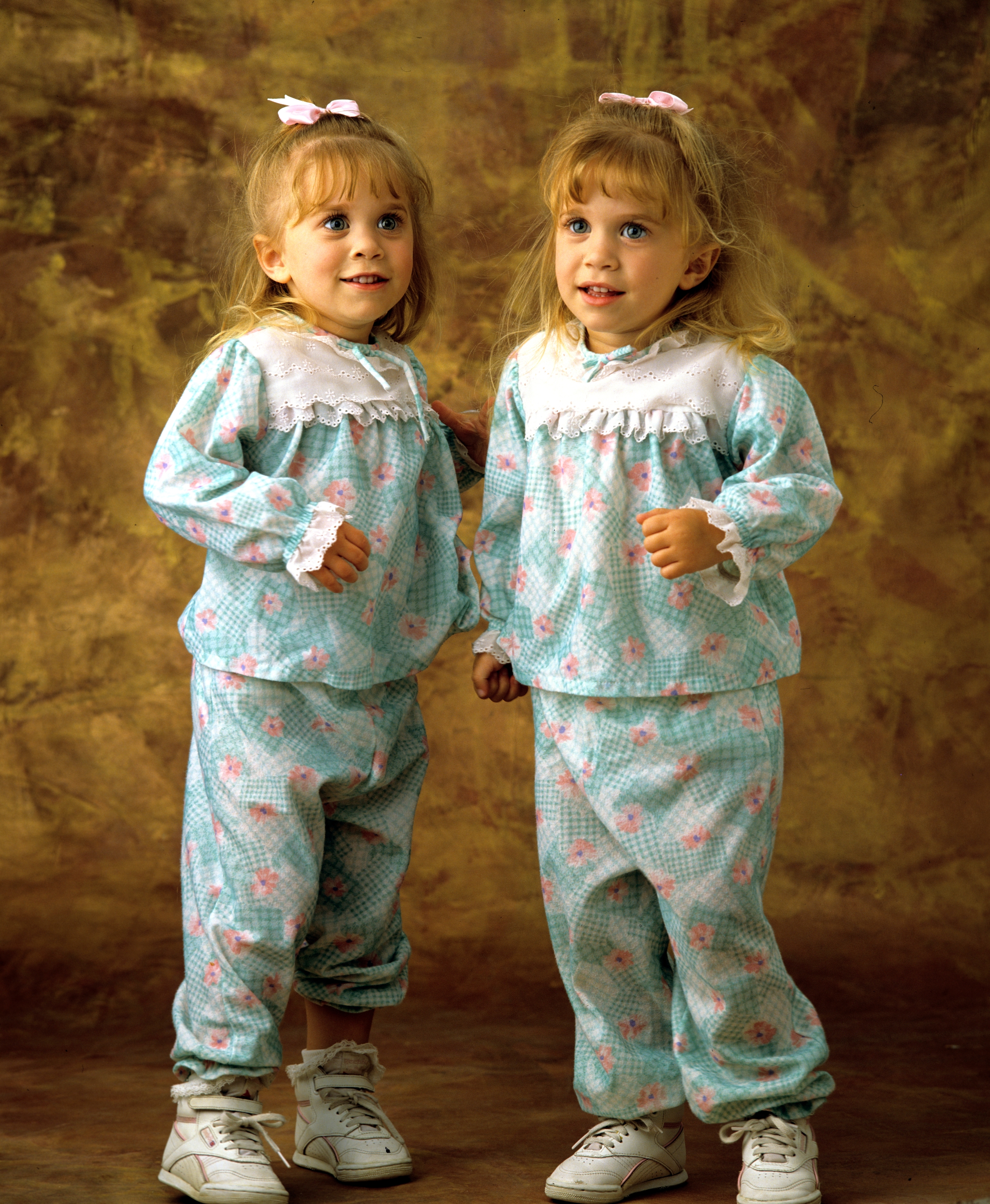 Two young girls in identical patterned outfits with lace collars and bows in their hair, standing together against a neutral background