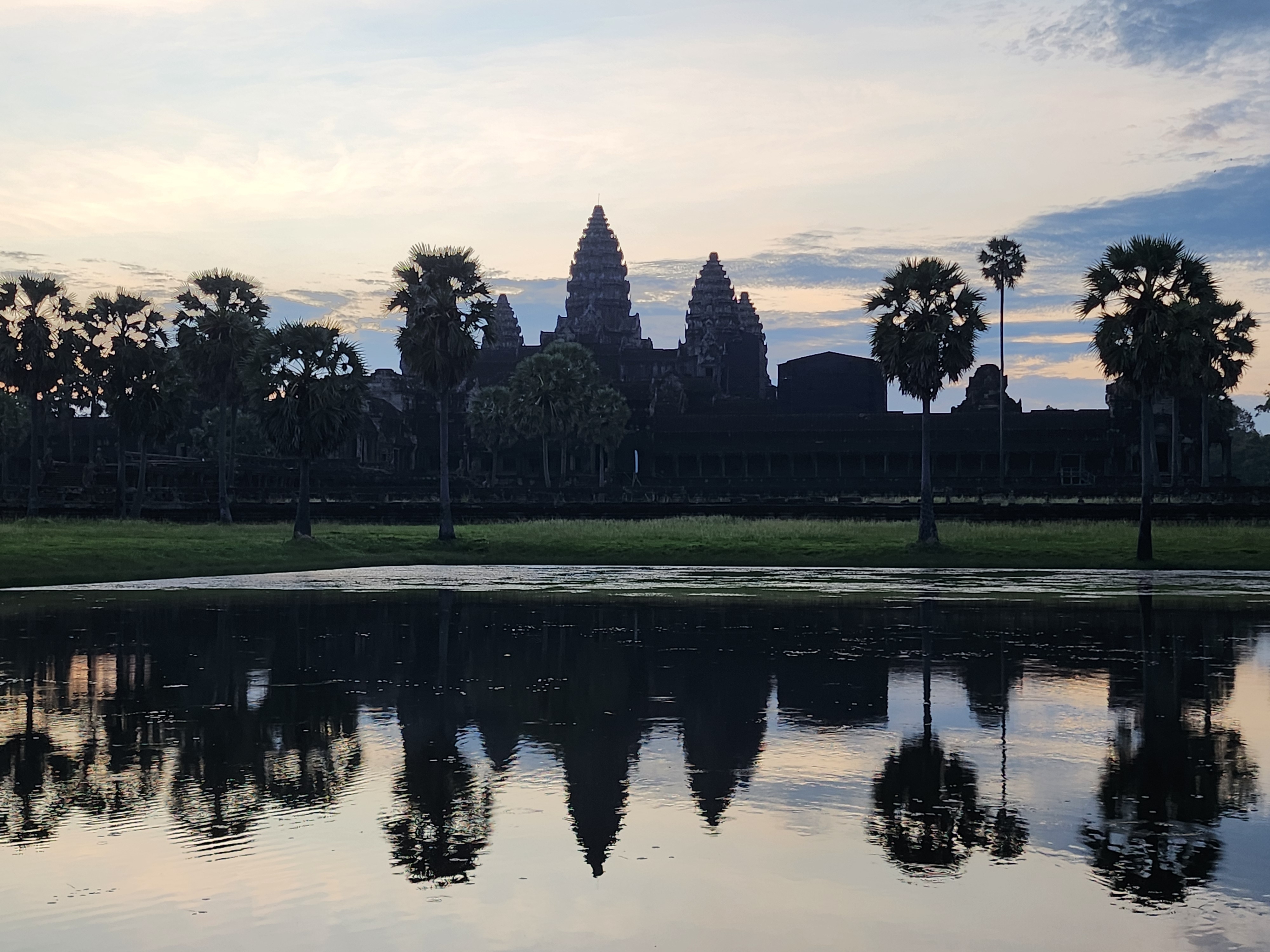 Angkor Wat temple at sunrise, reflected in a calm lake, with palm trees silhouetted in the foreground