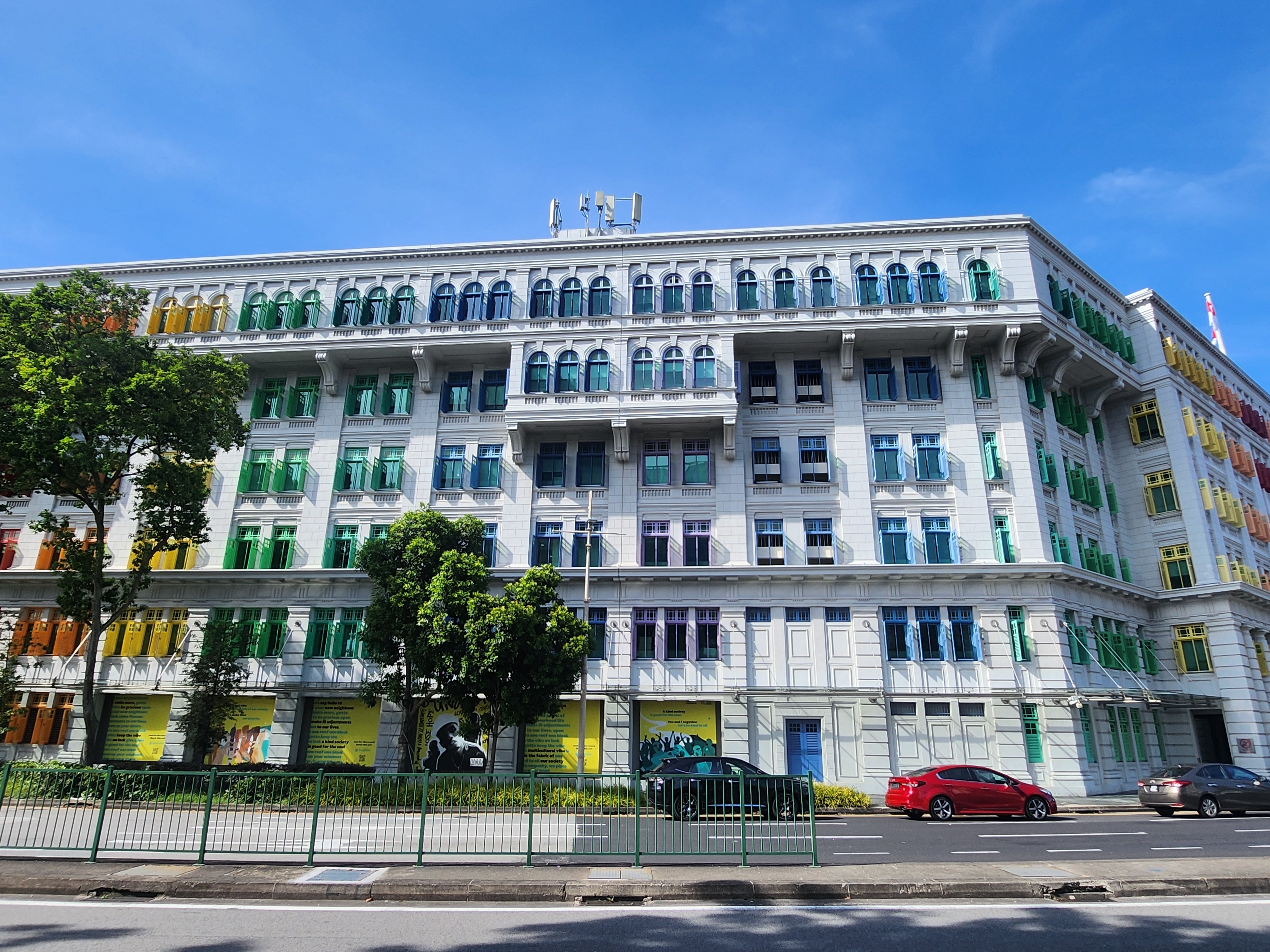Historic multi-story building with colorful window shutters and trees lining the sidewalk; two cars are parked in front