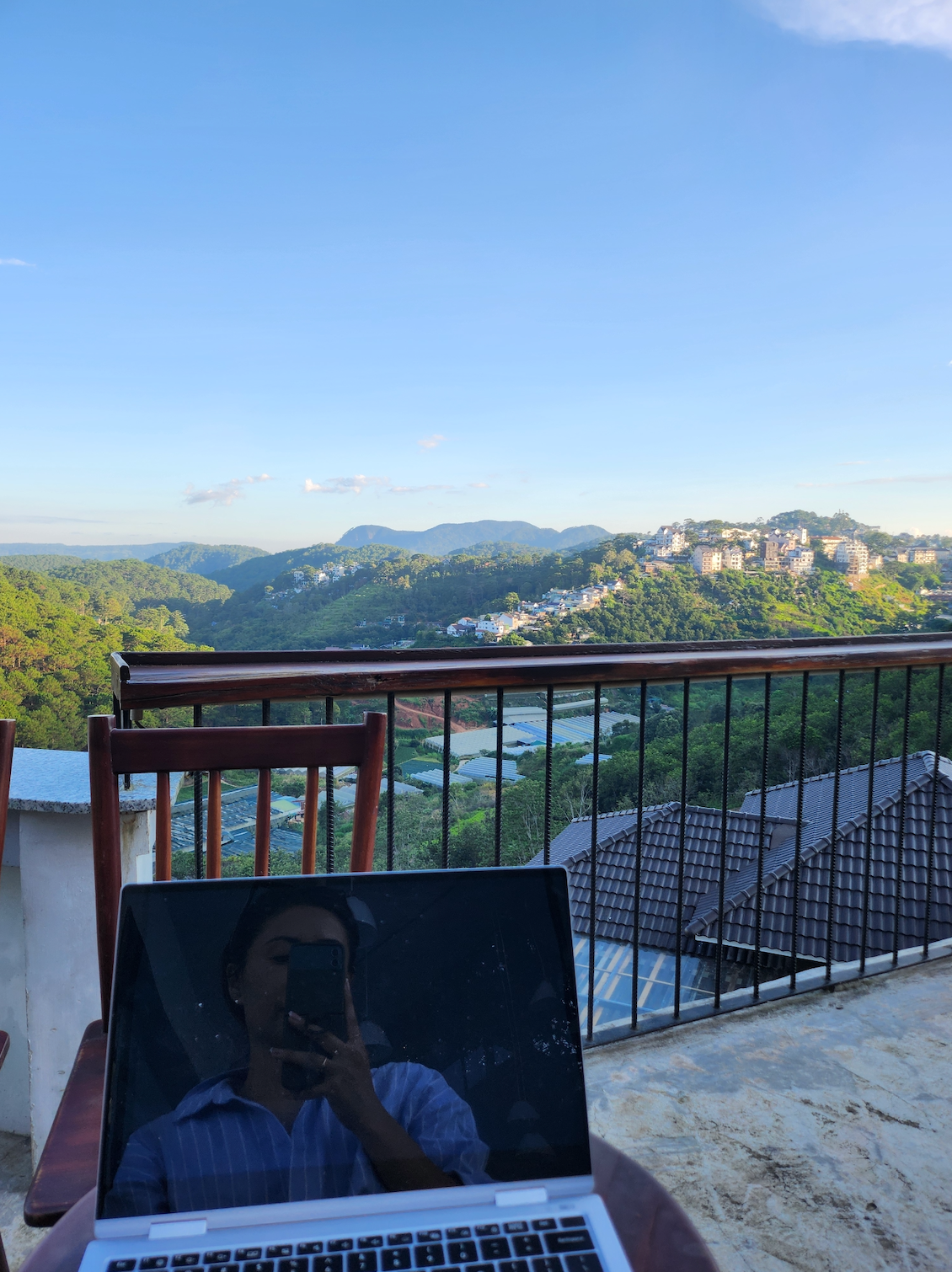 Author working on a laptop on a balcony with a scenic view of hills and houses, suggesting a remote work setup
