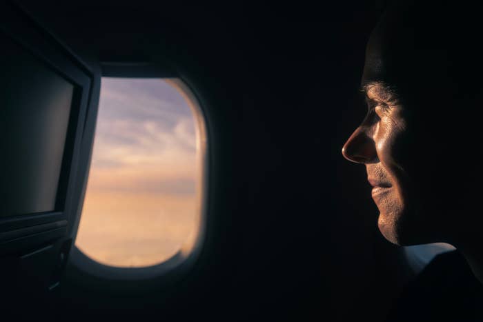 Person looking out an airplane window, with a screen beside them, suggesting travel during sunset