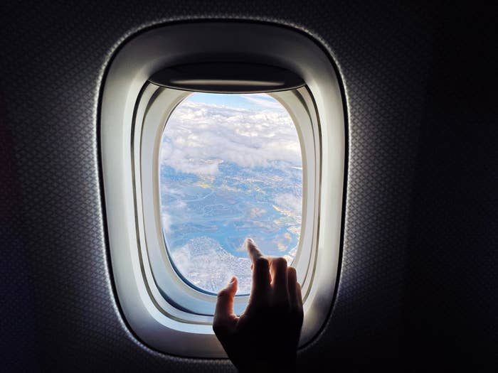 A hand points to a landscape view of clouds and land through an airplane window, indicating air travel