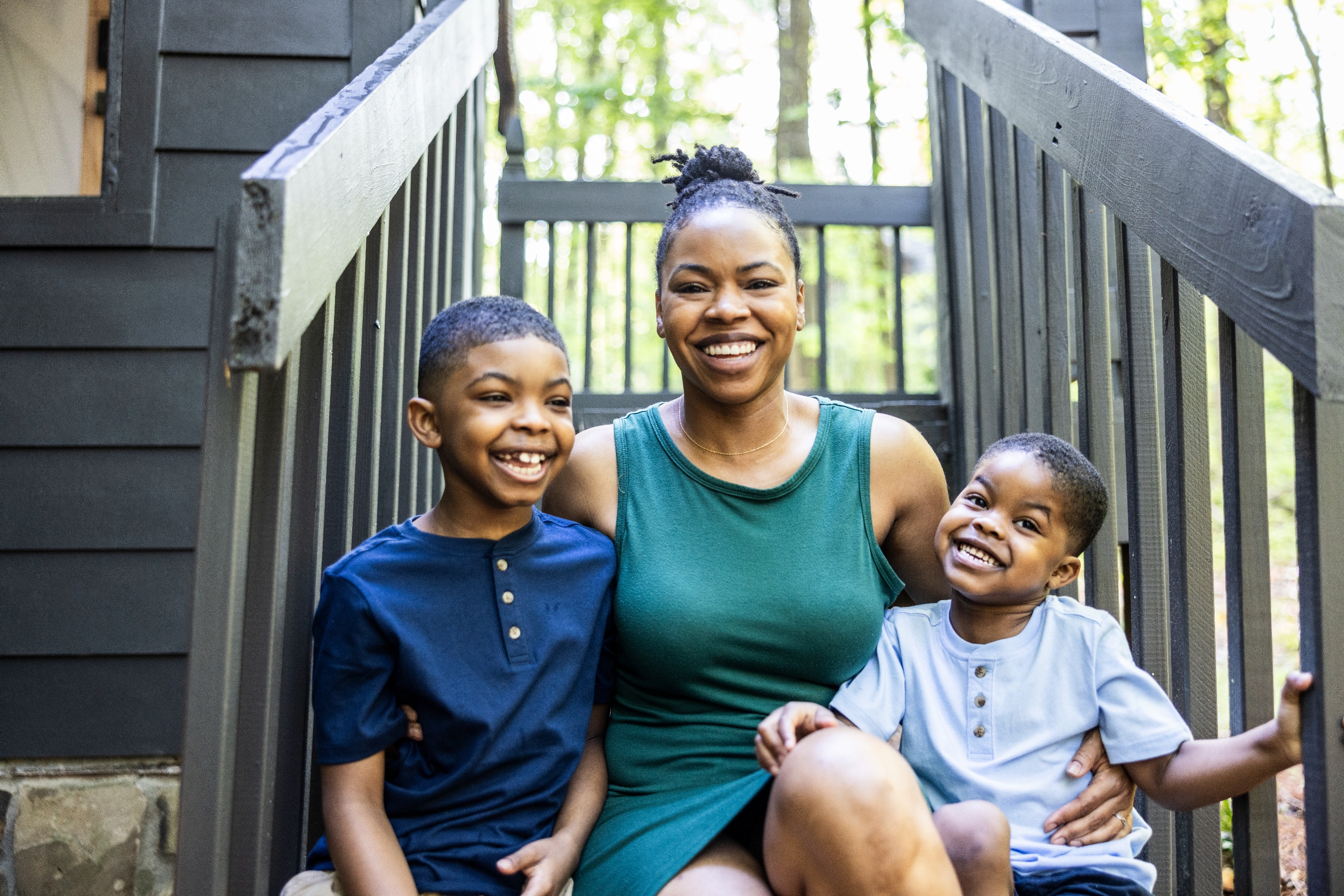 Smiling mother sits on outdoor steps with her two young sons, all casually dressed, surrounded by greenery