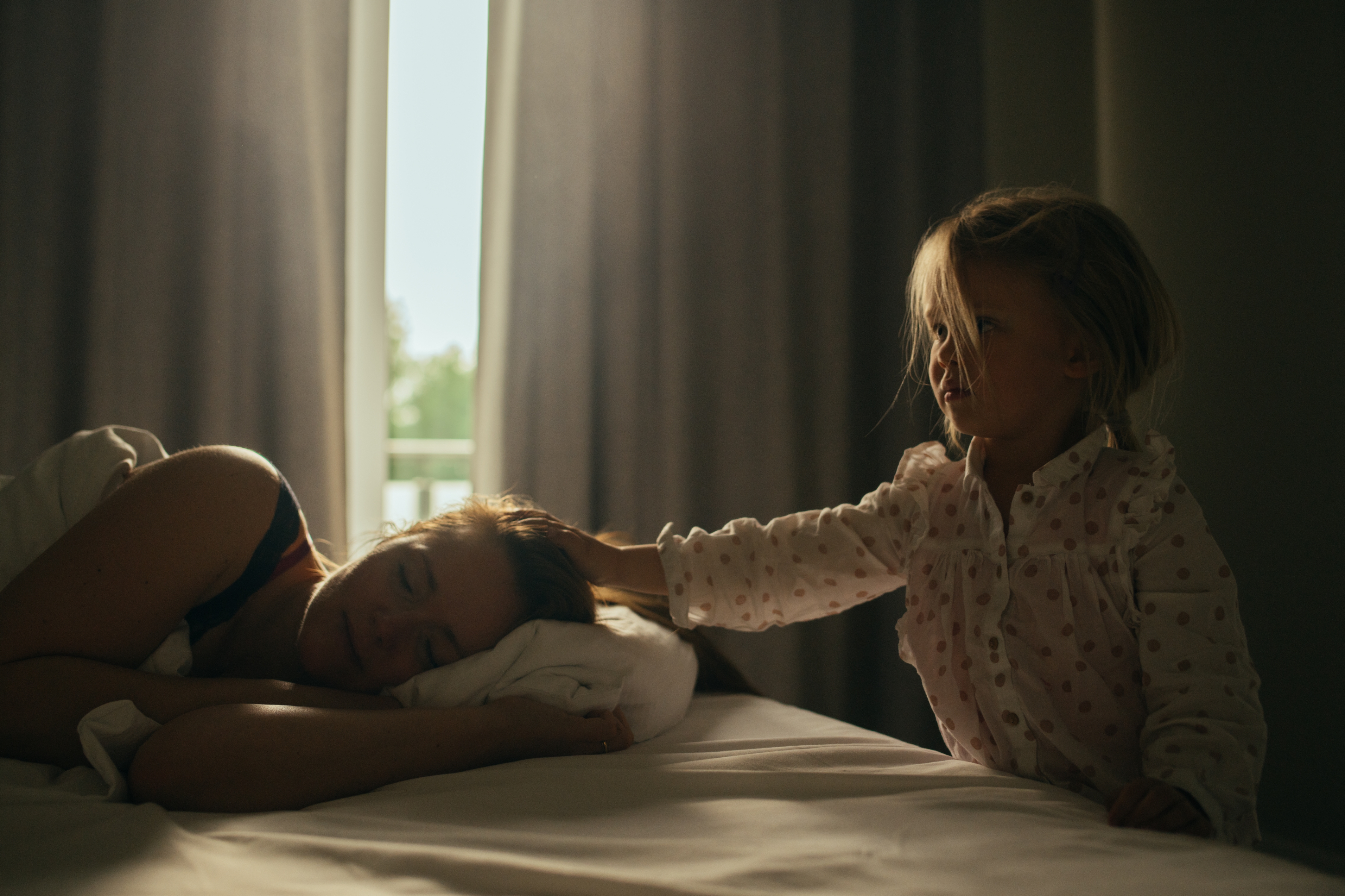 Child gently touches sleeping woman's hair in a cozy bedroom setting. Curtains softly filter light, creating a serene atmosphere