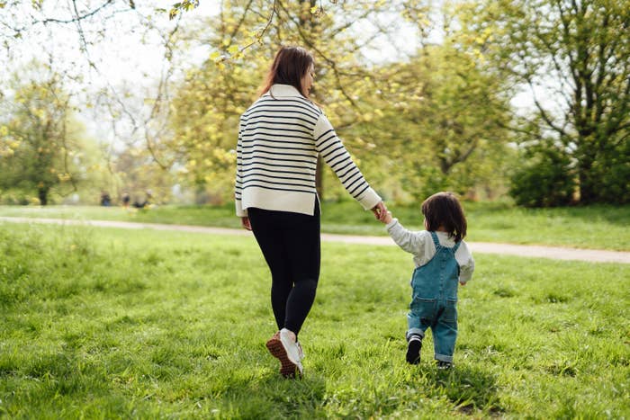 A woman and child holding hands walk in a park. The woman wears a striped sweater and leggings; the child wears overalls. Trees and grass surround them