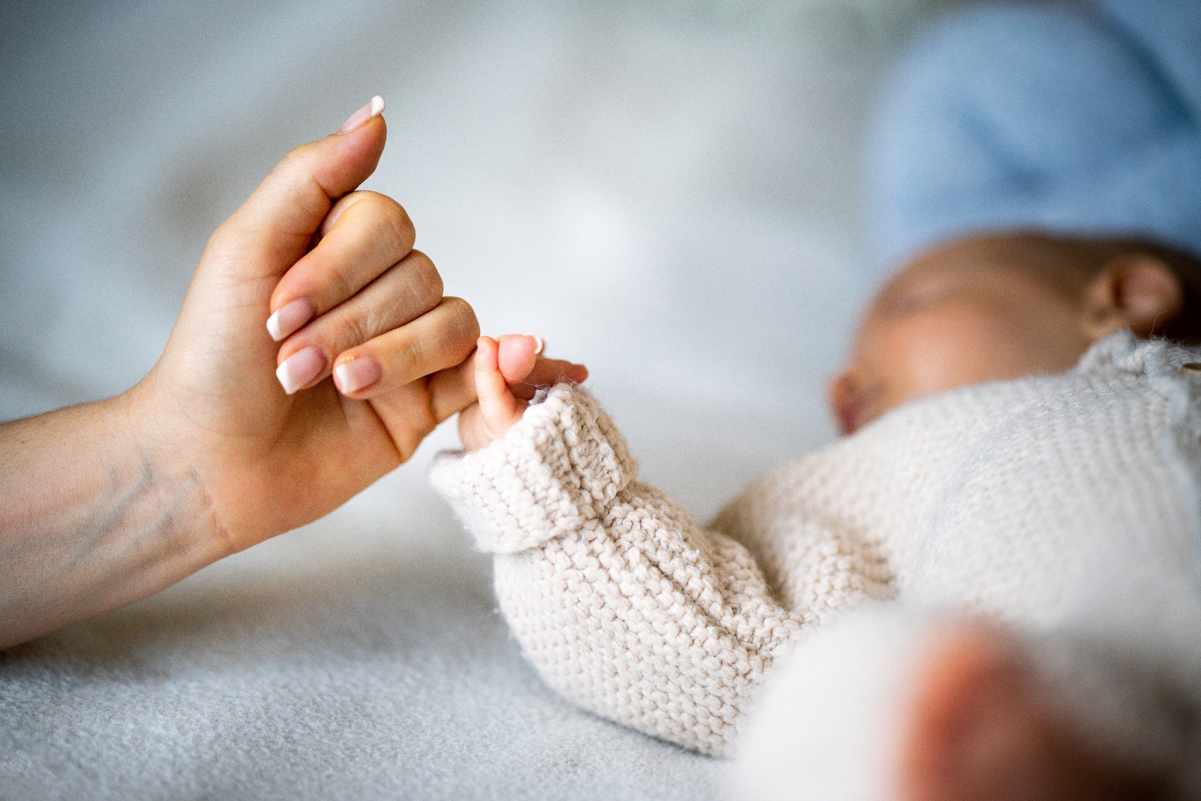Adult hand gently holding a baby's hand. The baby wears a knitted sweater and is lying down, creating a tender, parental moment