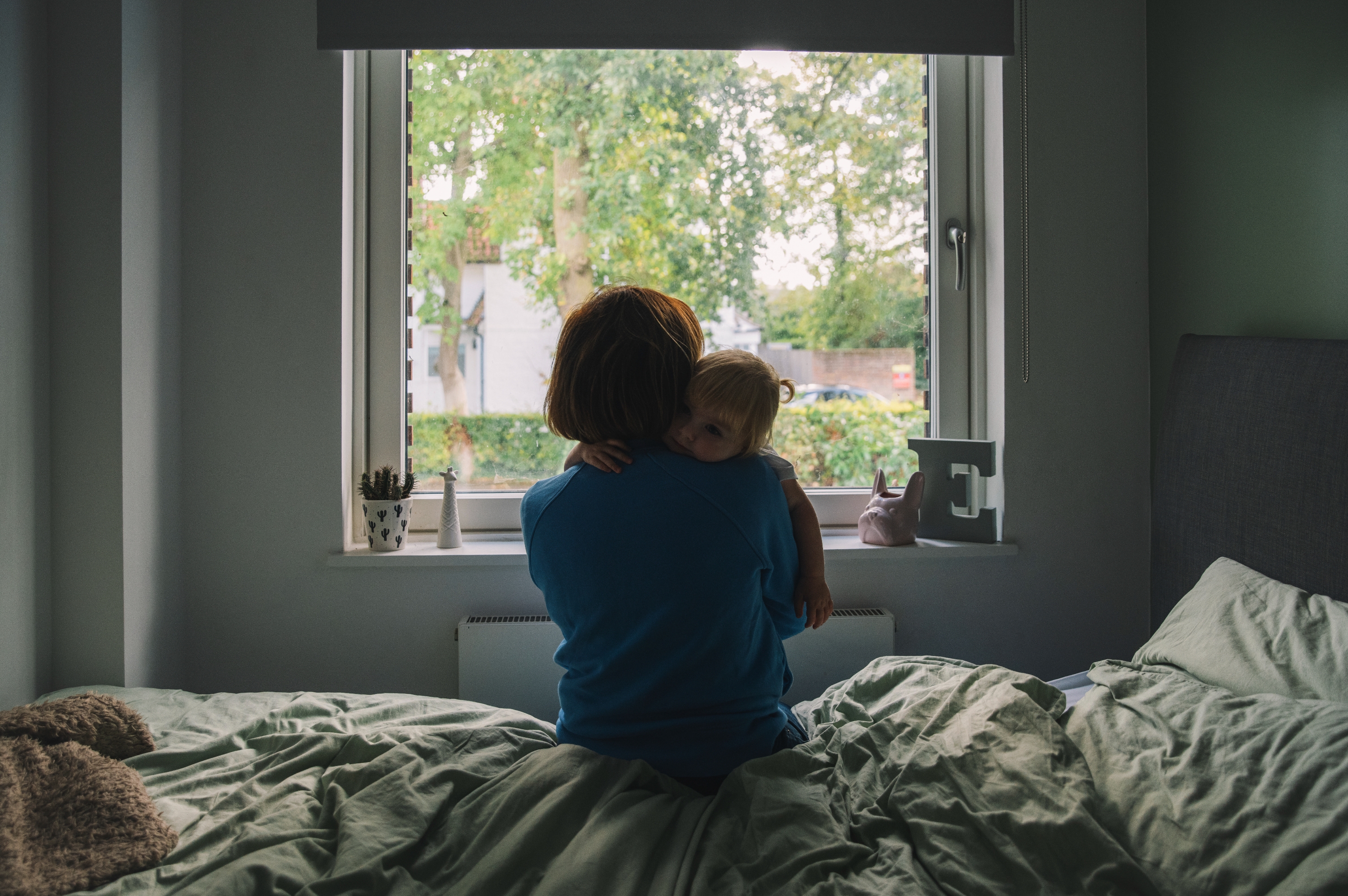 Person sitting on a bed, holding and comforting a young child near a window. The scene suggests a moment of gentle care and warmth