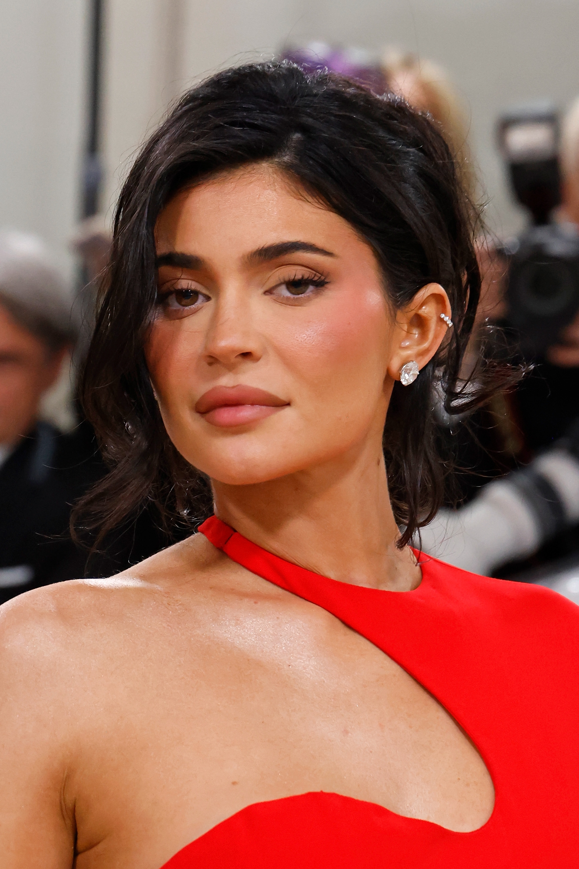 Person in elegant, asymmetrical dress poses on red carpet, with makeup accentuating features and hair styled in soft waves