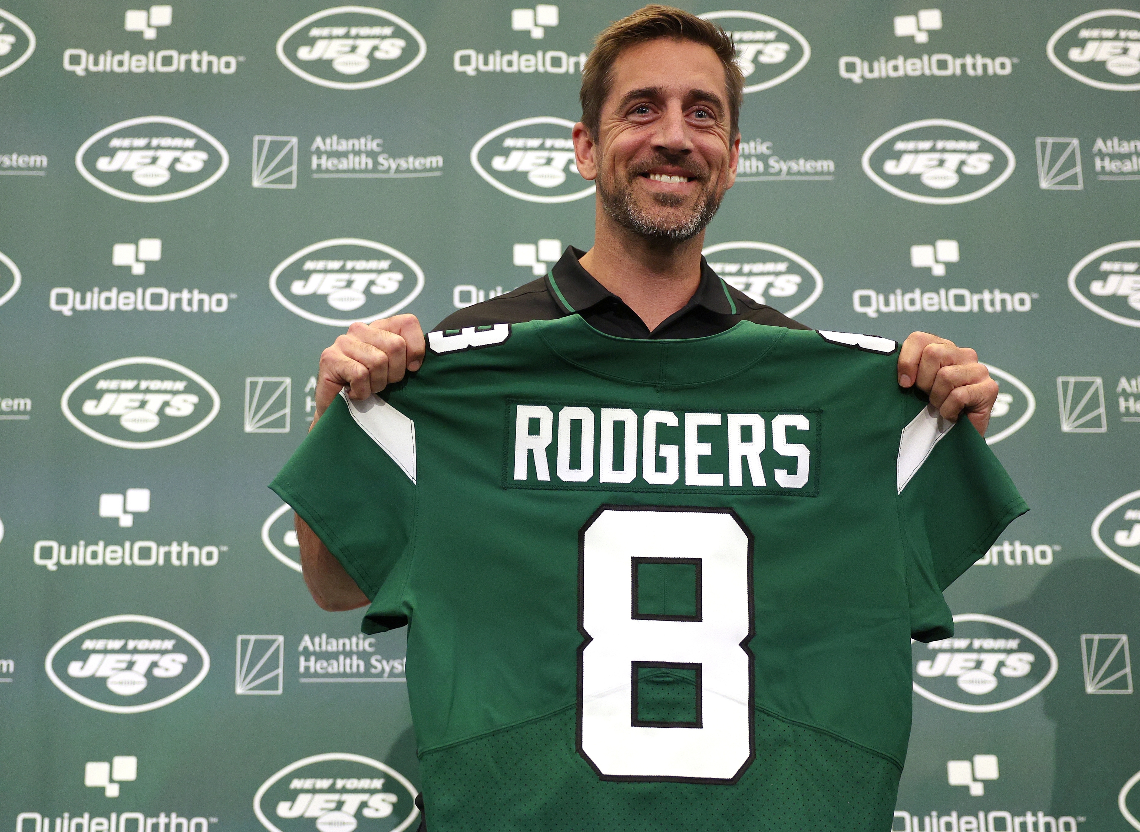 Person smiling, holding up a New York Jets jersey with the name "Rodgers" and number 8, at a sports team event backdrop