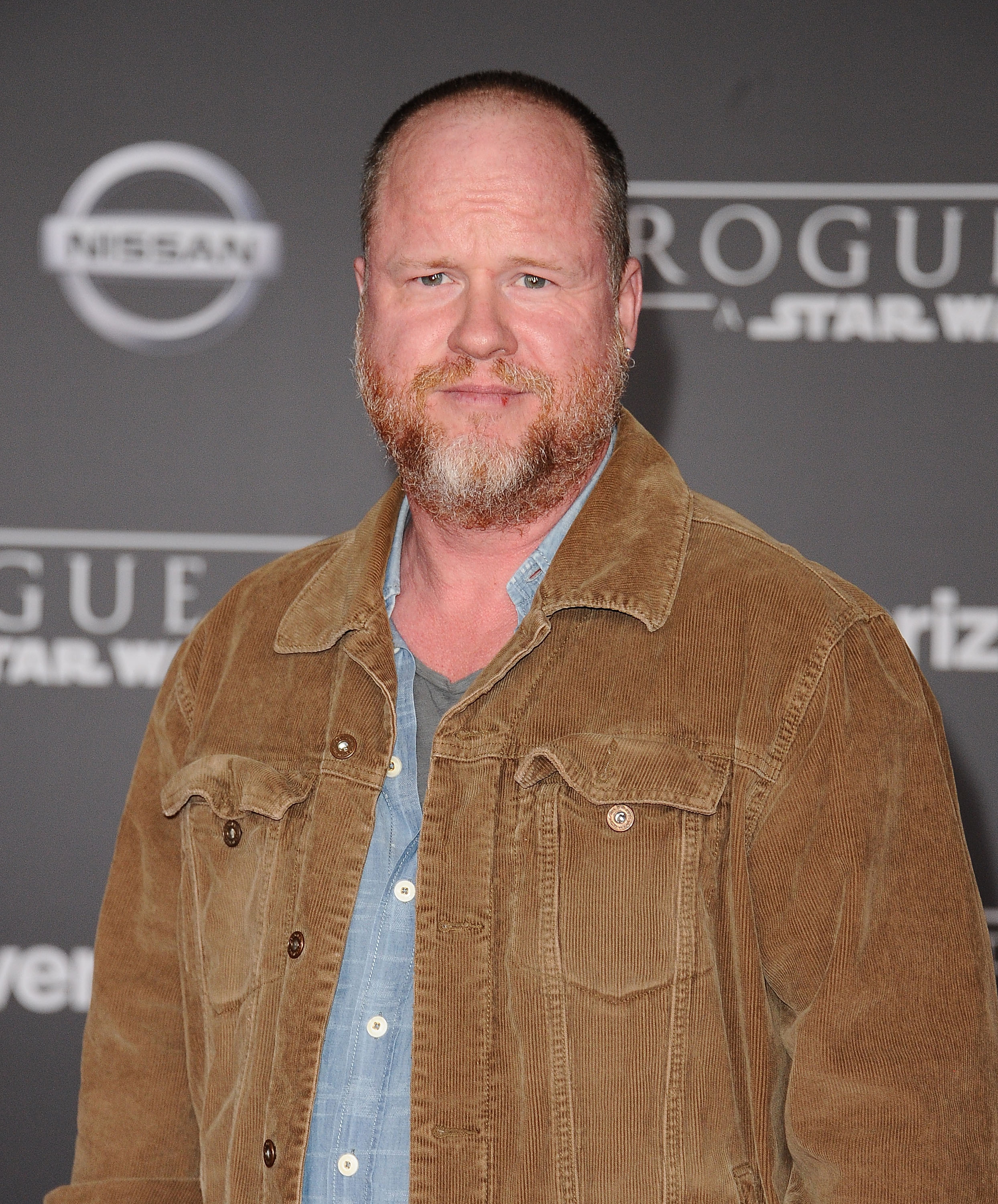 A person in a casual outfit with a jacket and shirt, standing on a media event backdrop