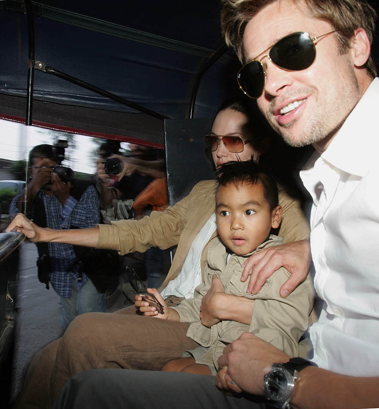 Two celebrities in casual attire are seated with a child on their lap, riding a tuk-tuk while photographers capture the moment