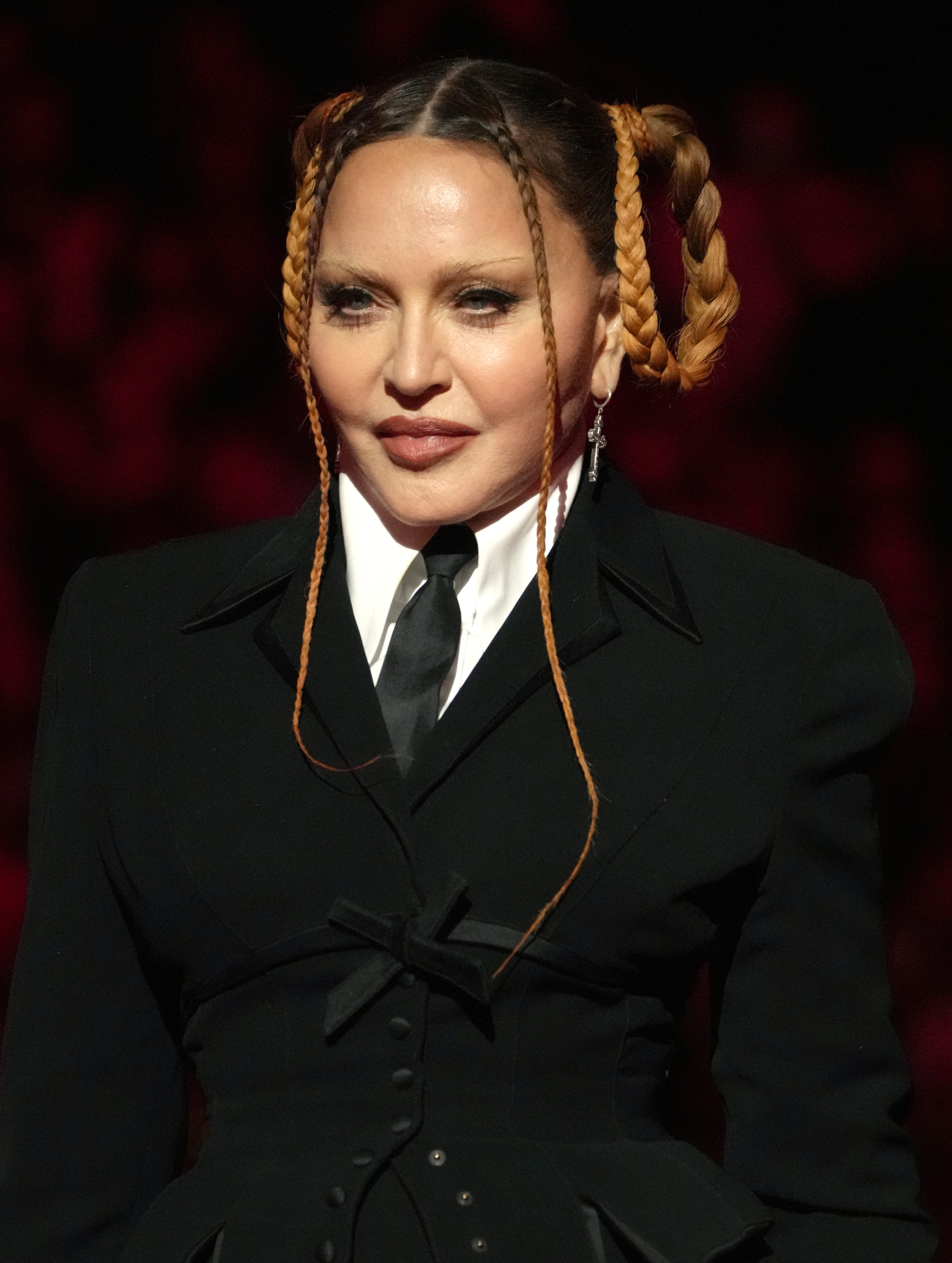 A person in a sleek, black suit with a tie and braided hairstyle, posing elegantly